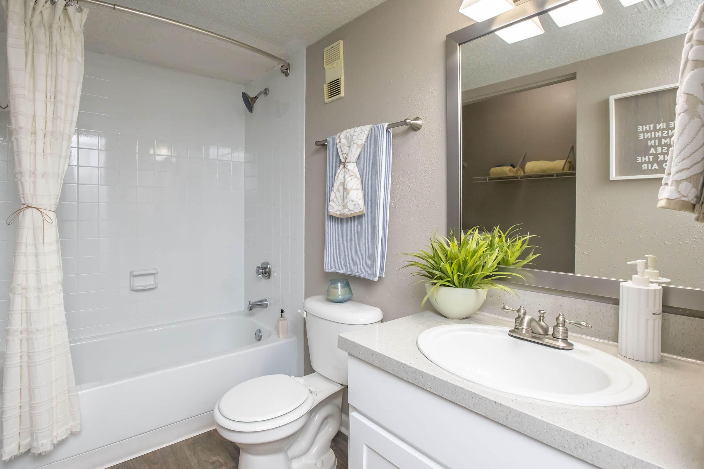 A bright and clean bathroom featuring a white bathtub with a shower, a toilet, a sleek countertop with a sink, and a decorative plant. The walls are painted in soft gray, and there are towels hanging on a rack. A mirror and light fixtures add to the modern aesthetic of the space.