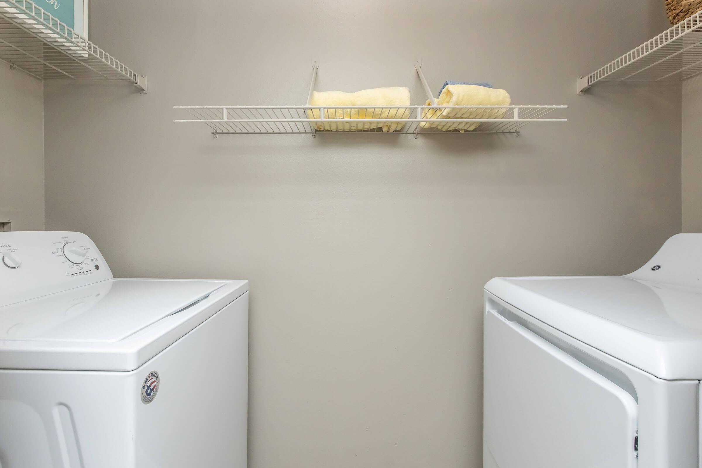 A laundry room featuring a white washing machine and dryer. Above them, a shelf holds neatly stacked towels. The walls are a neutral gray, providing a clean and organized appearance.