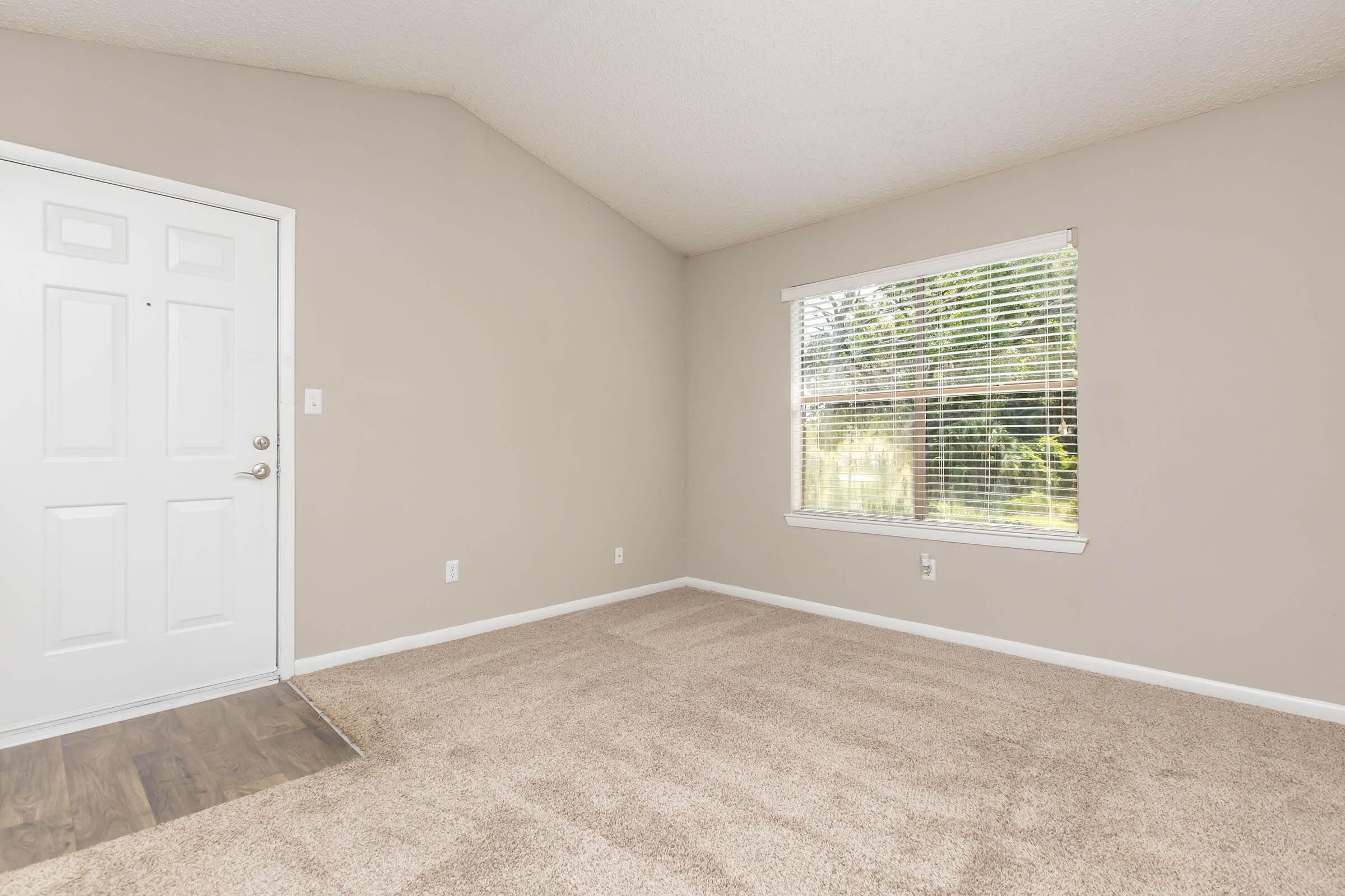 A spacious room with light brown carpet, featuring a white front door on the left and a large window with white blinds allowing natural light in. The walls are painted a soft beige, and there are no furnishings, creating an empty and inviting atmosphere.