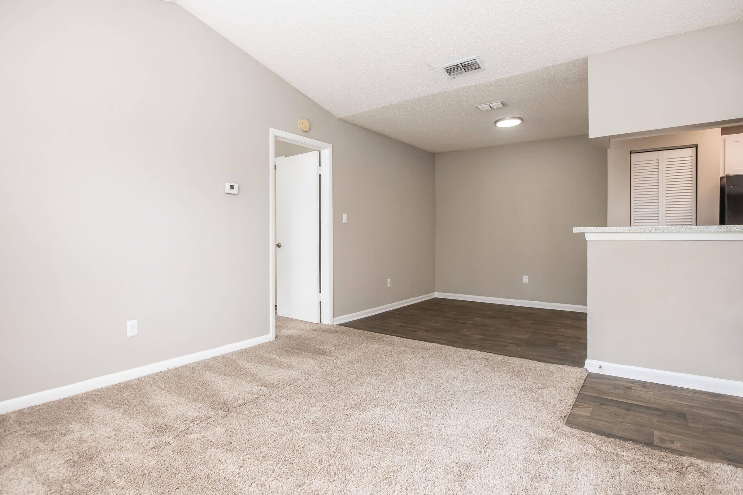 A spacious, neutrally colored living area featuring carpet flooring and a small kitchen area in the background. There is a white door leading to another room and a light fixture in the ceiling. The walls are plain, enhancing the open feel of the space.