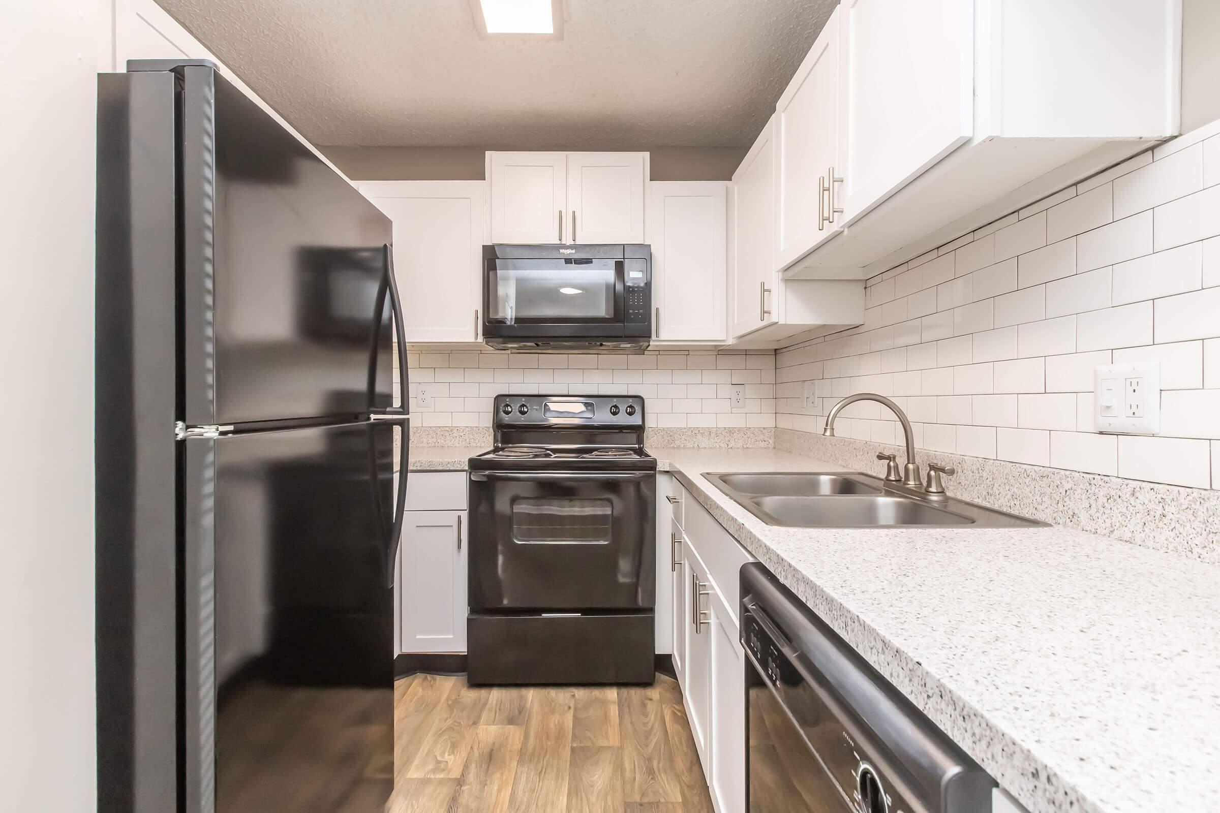 A modern kitchen featuring a black refrigerator, black oven, and microwave, all against white cabinets and a gray tiled backsplash. The countertops are light-colored granite, and there are dual sinks with a faucet. The floor is a wood-like laminate, creating a cozy atmosphere.