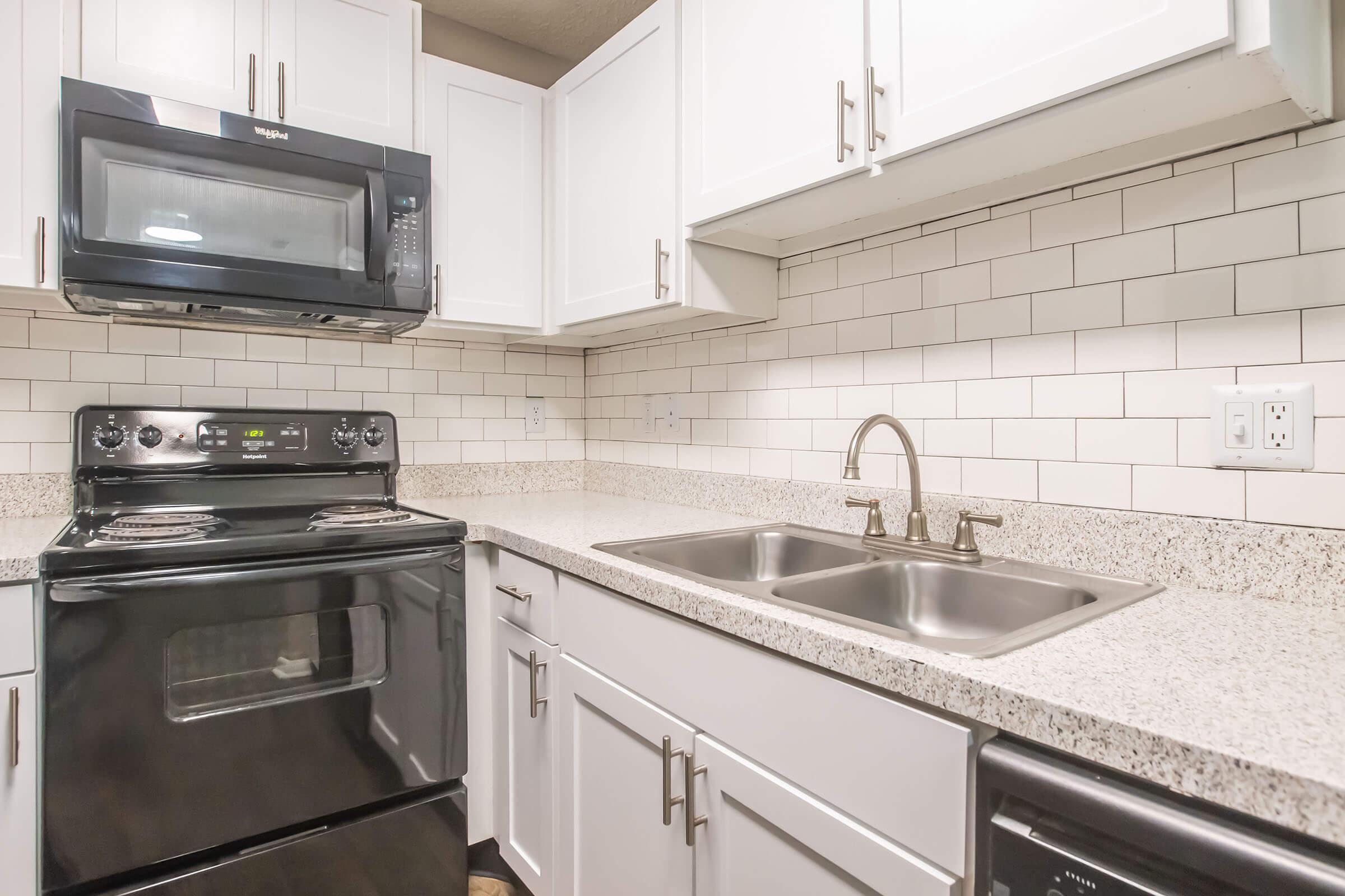 A modern kitchen featuring a black stove and microwave, white cabinetry, and a granite countertop. There are dual stainless steel sinks with a brushed nickel faucet and a textured tile backsplash. In the corner, an under-counter refrigerator is visible.