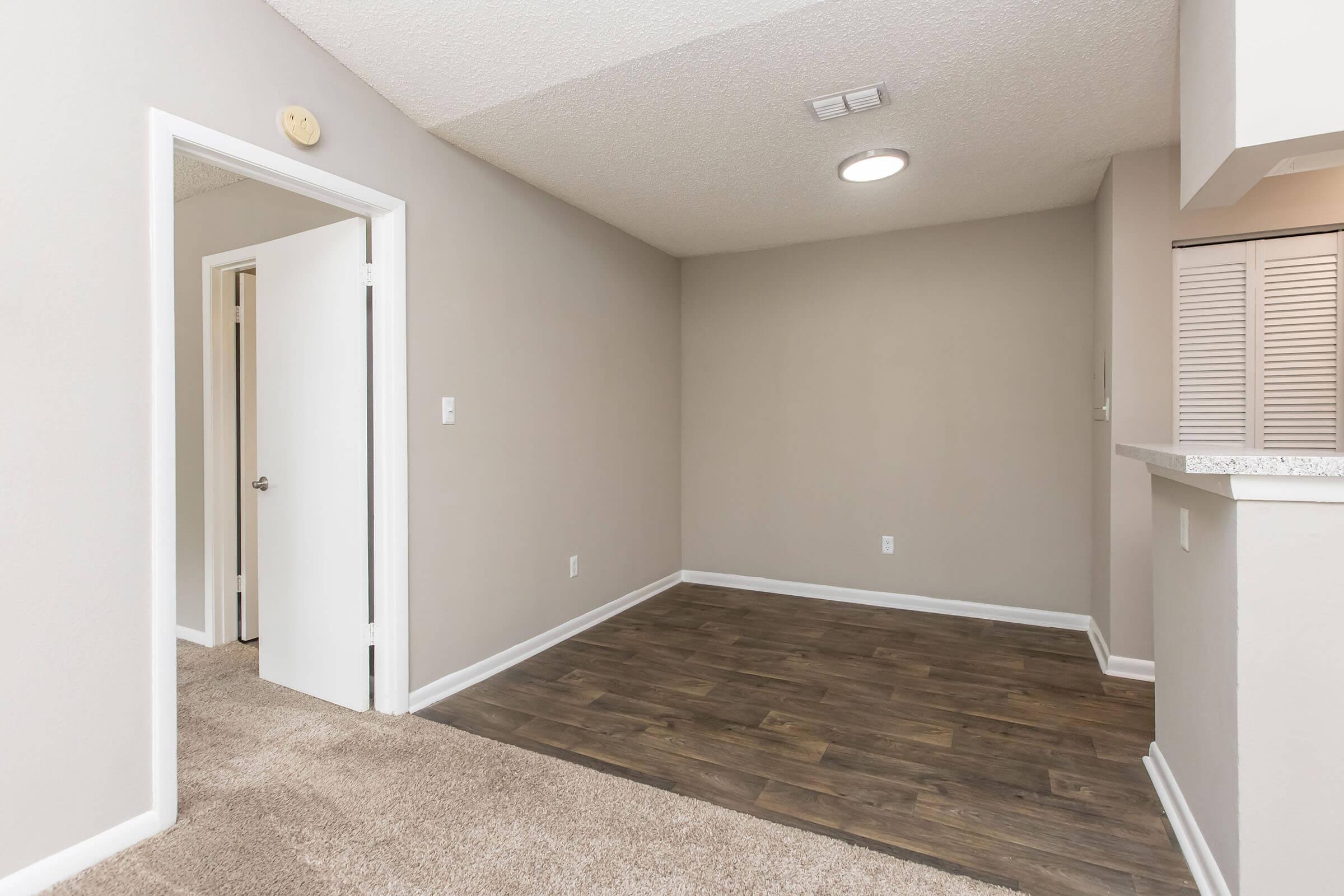Empty room featuring beige walls and a textured ceiling. There is a light fixture on the ceiling, a doorway leading to another room on the left, and a corner with a small bar or counter area. The floor is covered with wood-like laminate, and there is a carpeted area in the foreground.
