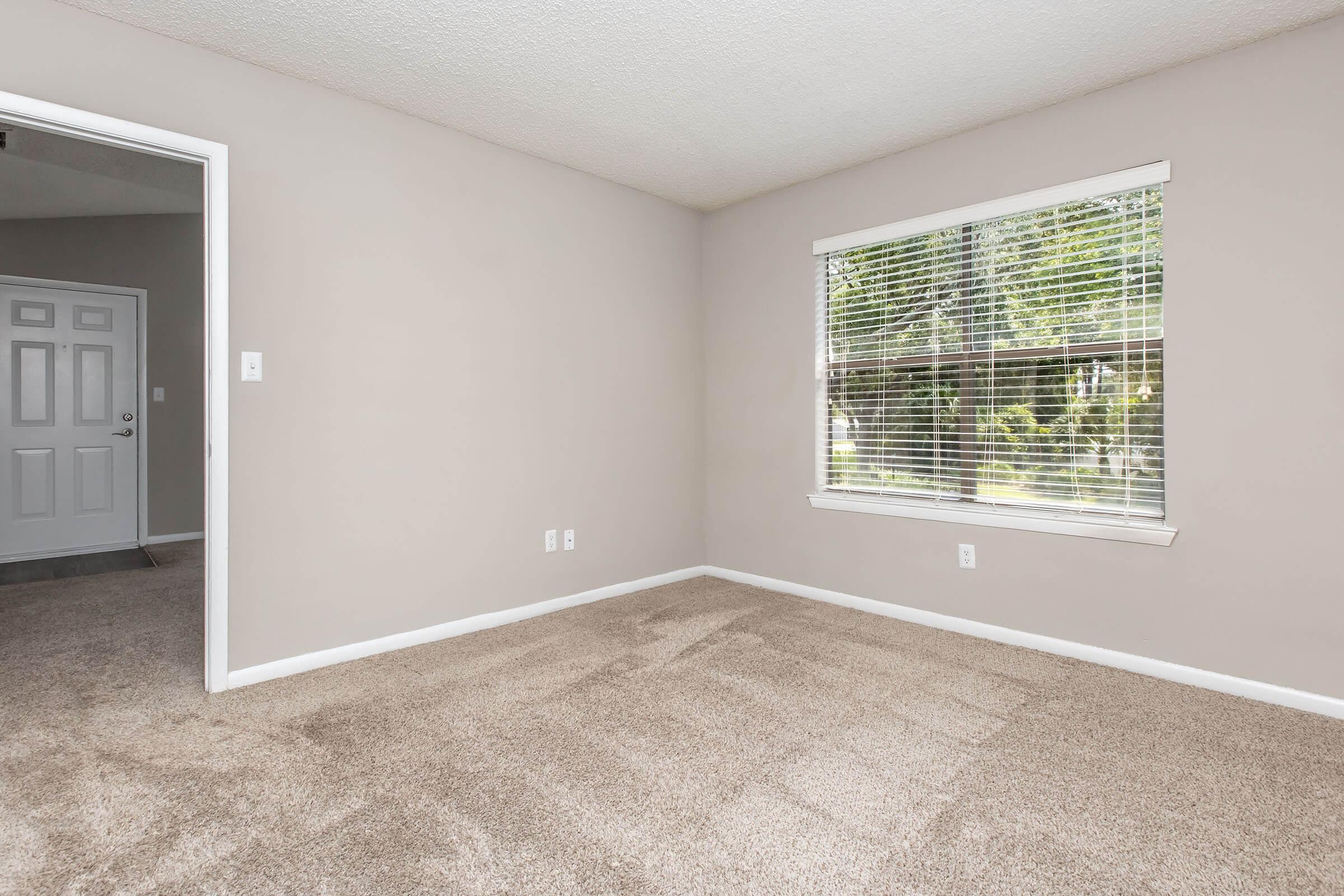 A vacant room featuring beige walls and a large window with white blinds, letting in natural light. The floor is covered with light brown carpet. A door can be seen on the left side, leading to another space. The room is empty, creating a spacious and clean appearance ideal for various uses.