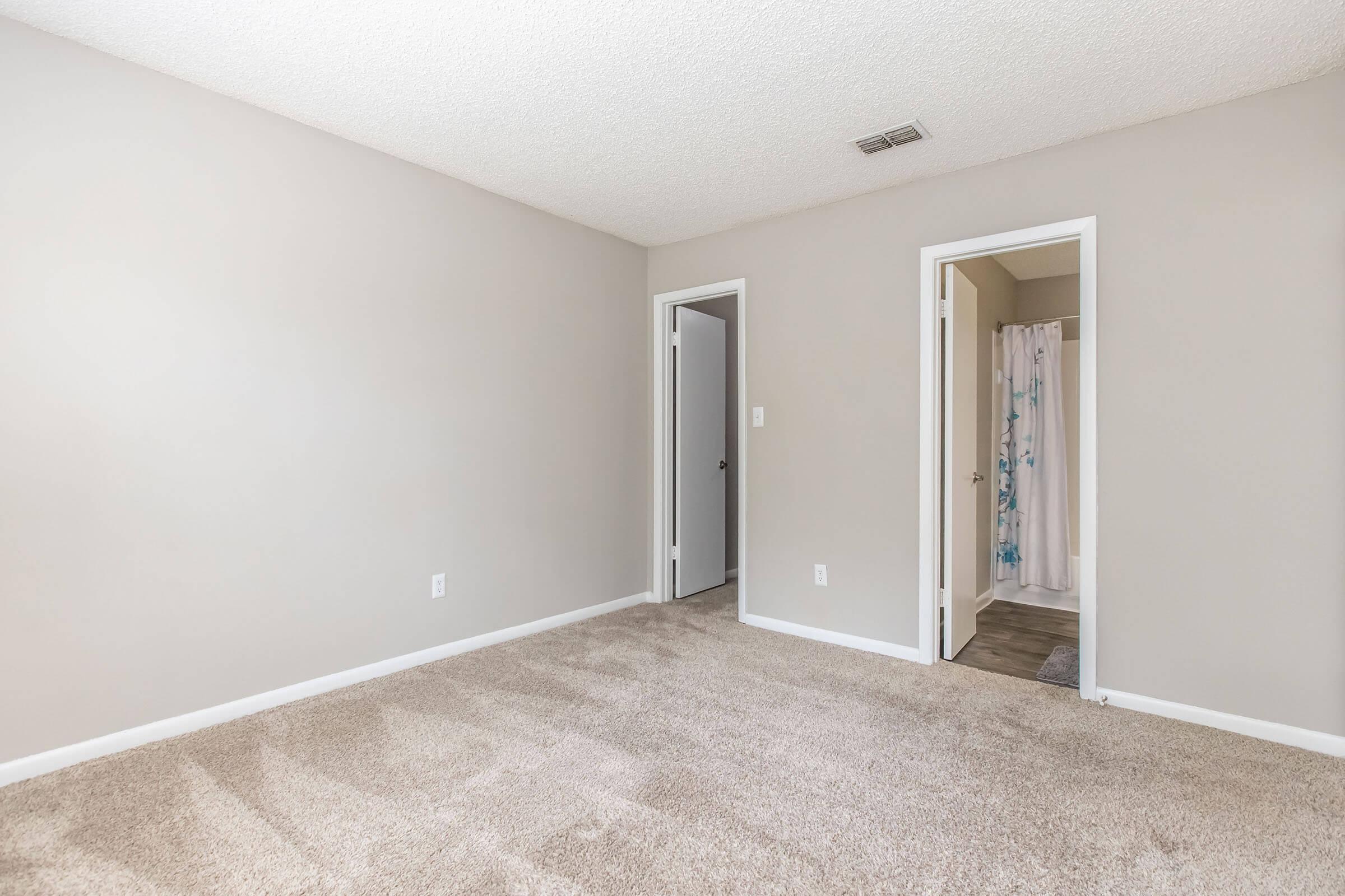 A spacious, empty room with light gray walls and beige carpeting. There are two doorways, one leading to a bathroom with a shower curtain featuring a blue design. The room has a bright and airy feel, illuminated by natural light. The ceiling has a textured finish, adding to the overall aesthetic.