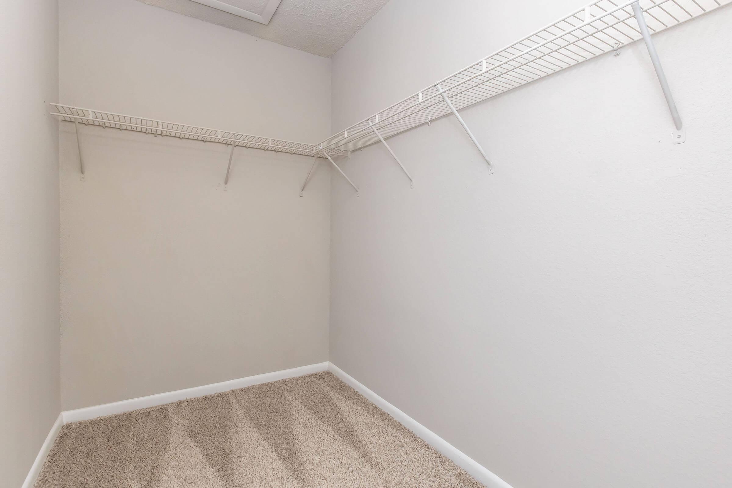 Empty closet with white walls and a light brown carpeted floor. There are wire shelves mounted on the walls, providing storage space. The room appears well-lit and clean, with no items or decorations visible.