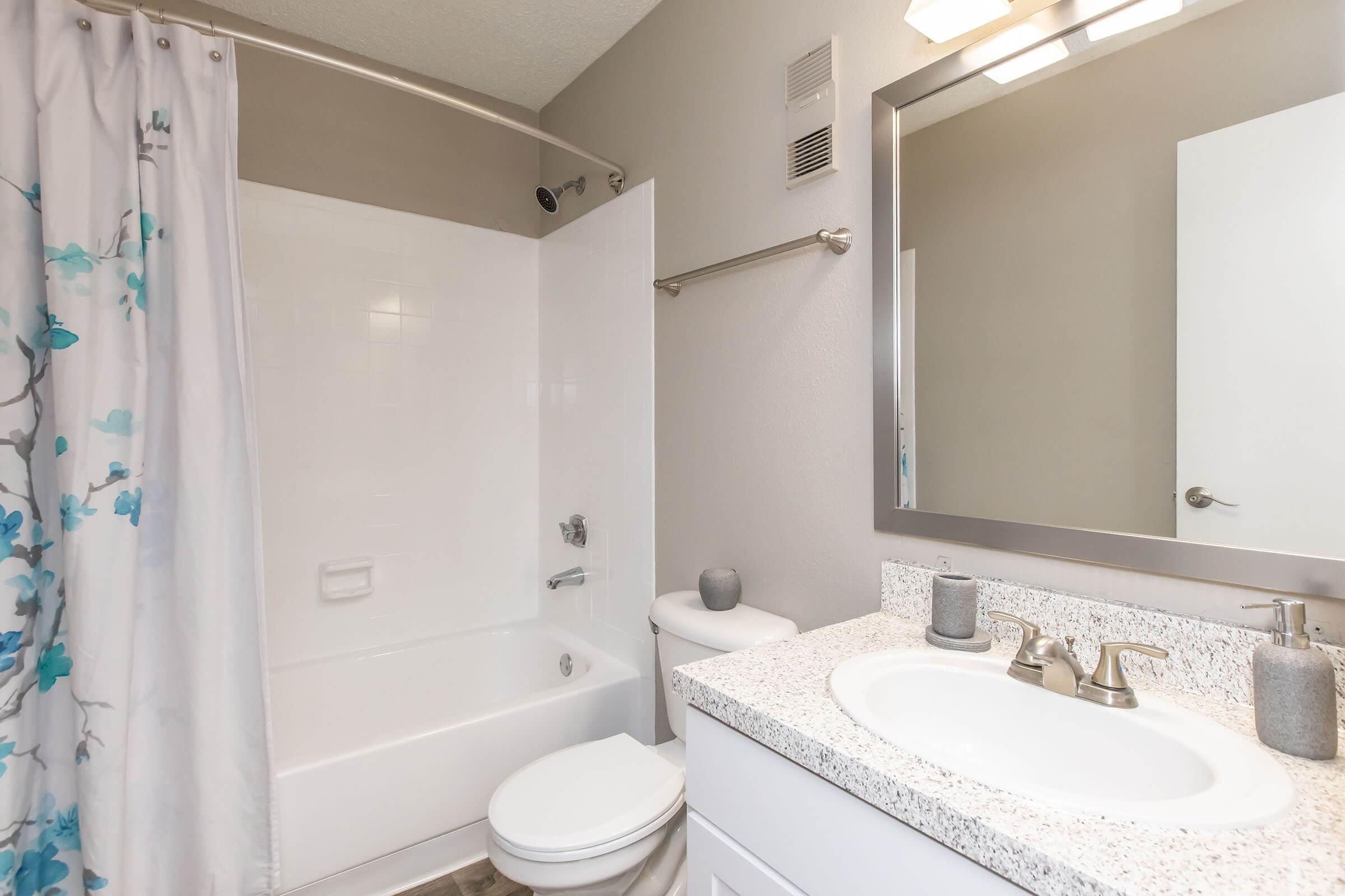 A modern bathroom featuring a white bathtub with a shower, a toilet, and a sink with a granite countertop. The shower curtain has a floral design in blue. The room has neutral-colored walls and a large mirror above the sink, providing a clean and bright atmosphere.