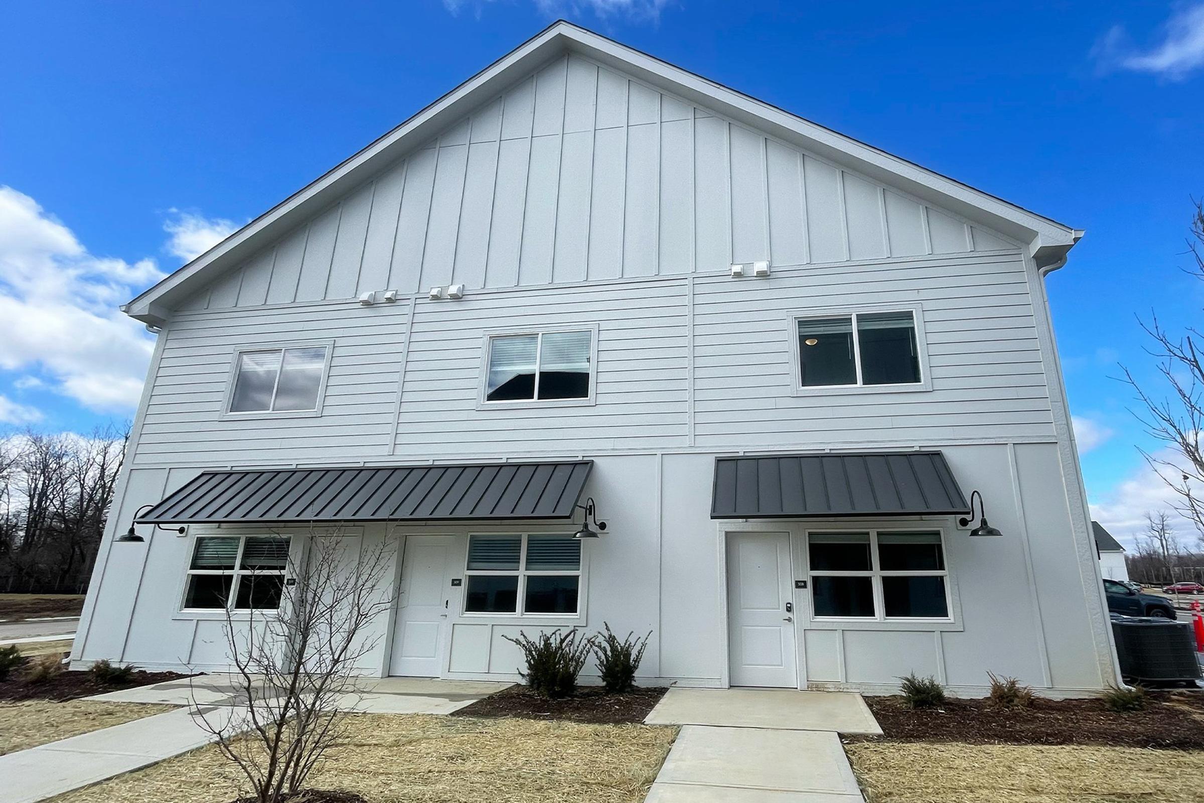 A modern two-story white building with a sloped roof, featuring two front entrances with black awnings and large windows. The surrounding area has neatly maintained landscaping, including small shrubs and grass, under a partially cloudy blue sky.