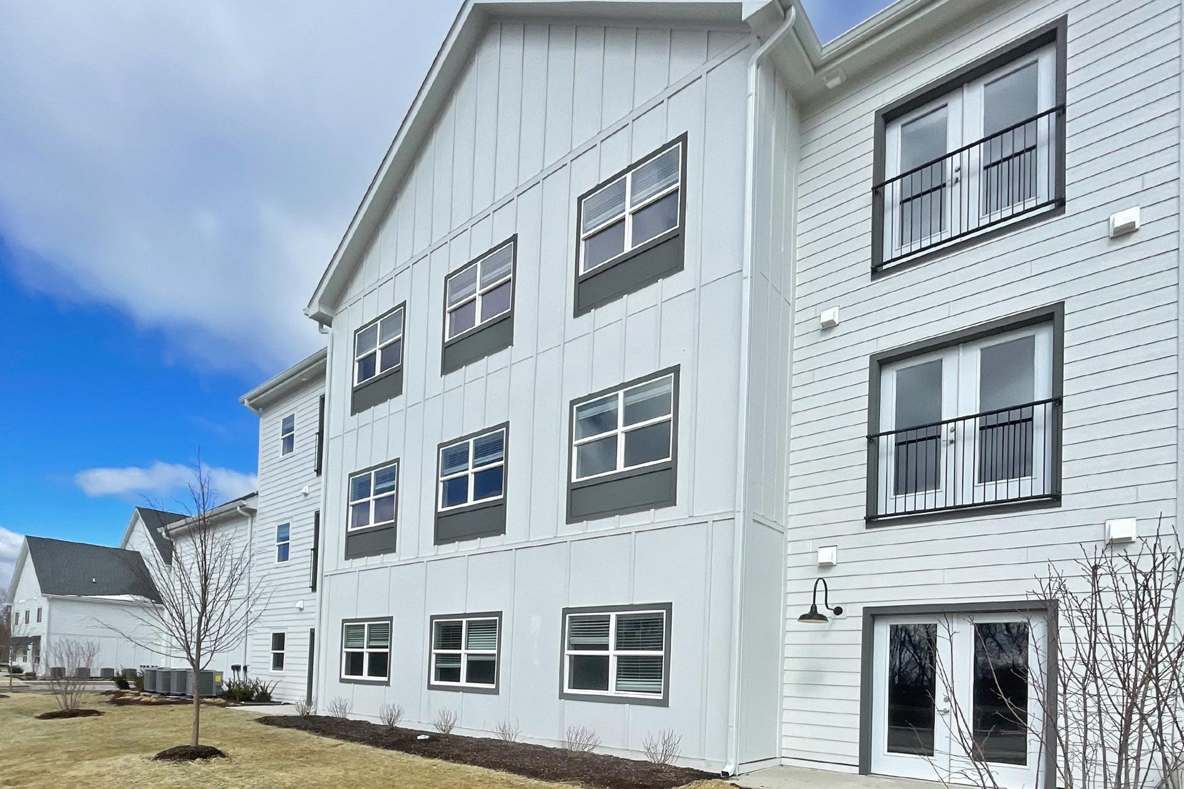 A modern, multi-story residential building with a white exterior and large windows. The ground area is landscaped with minimal vegetation, and the sky is partly cloudy. The building has a contemporary architectural style, featuring multiple balconies and a clean design.
