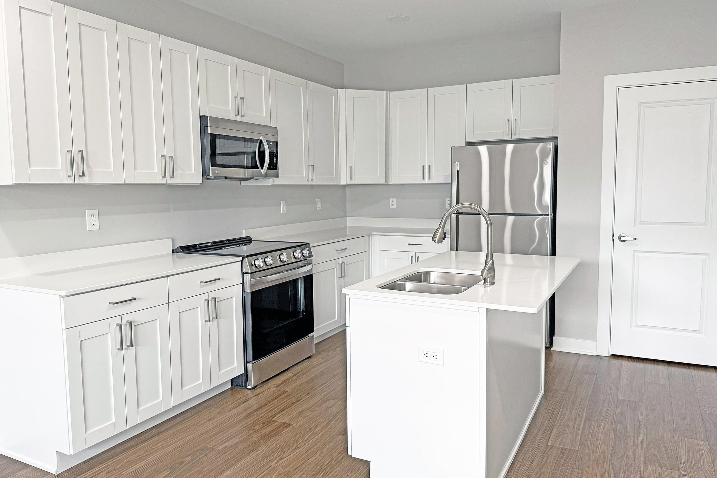 Modern kitchen featuring white cabinetry, stainless steel appliances including a refrigerator and oven, and a large island with a sink. The space has light-colored walls and wooden flooring, creating a bright and airy atmosphere.