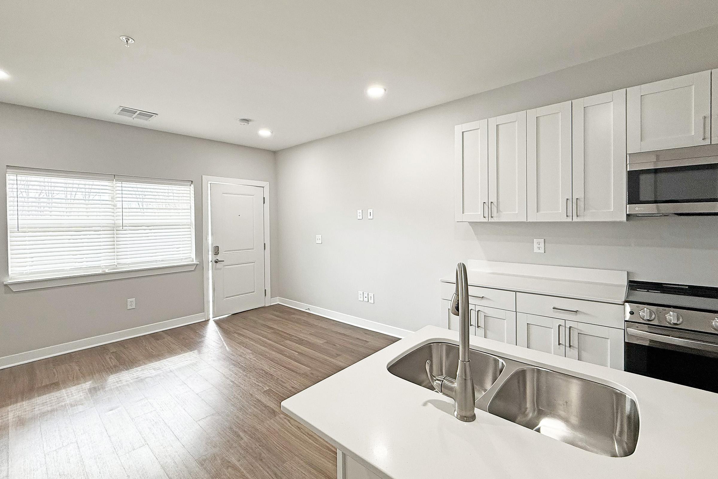 A modern kitchen and living area featuring a stainless steel sink, white cabinetry, and an adjacent door leading outside. The space has hardwood flooring, large windows with natural light, and a sleek stove and microwave in the kitchen, creating a bright and inviting atmosphere.