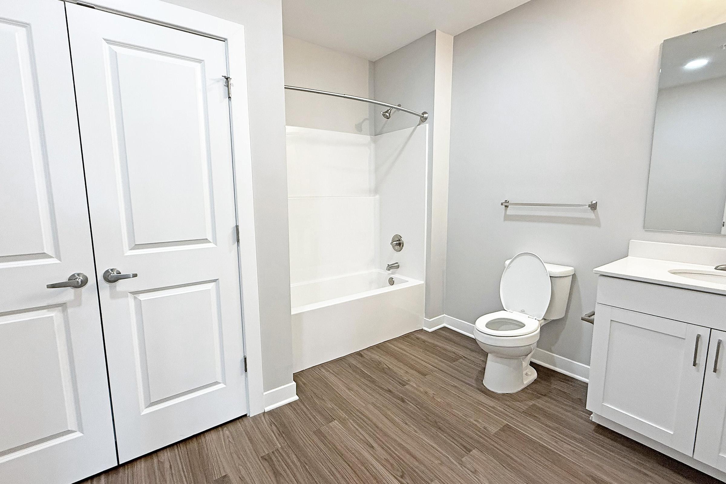 A modern bathroom featuring a white bathtub with a shower curtain rod, a toilet, and a vanity with a mirror. The walls are painted light gray, and the floor has wood-like tiles. There are double doors leading to another area, adding a spacious feel to the room.