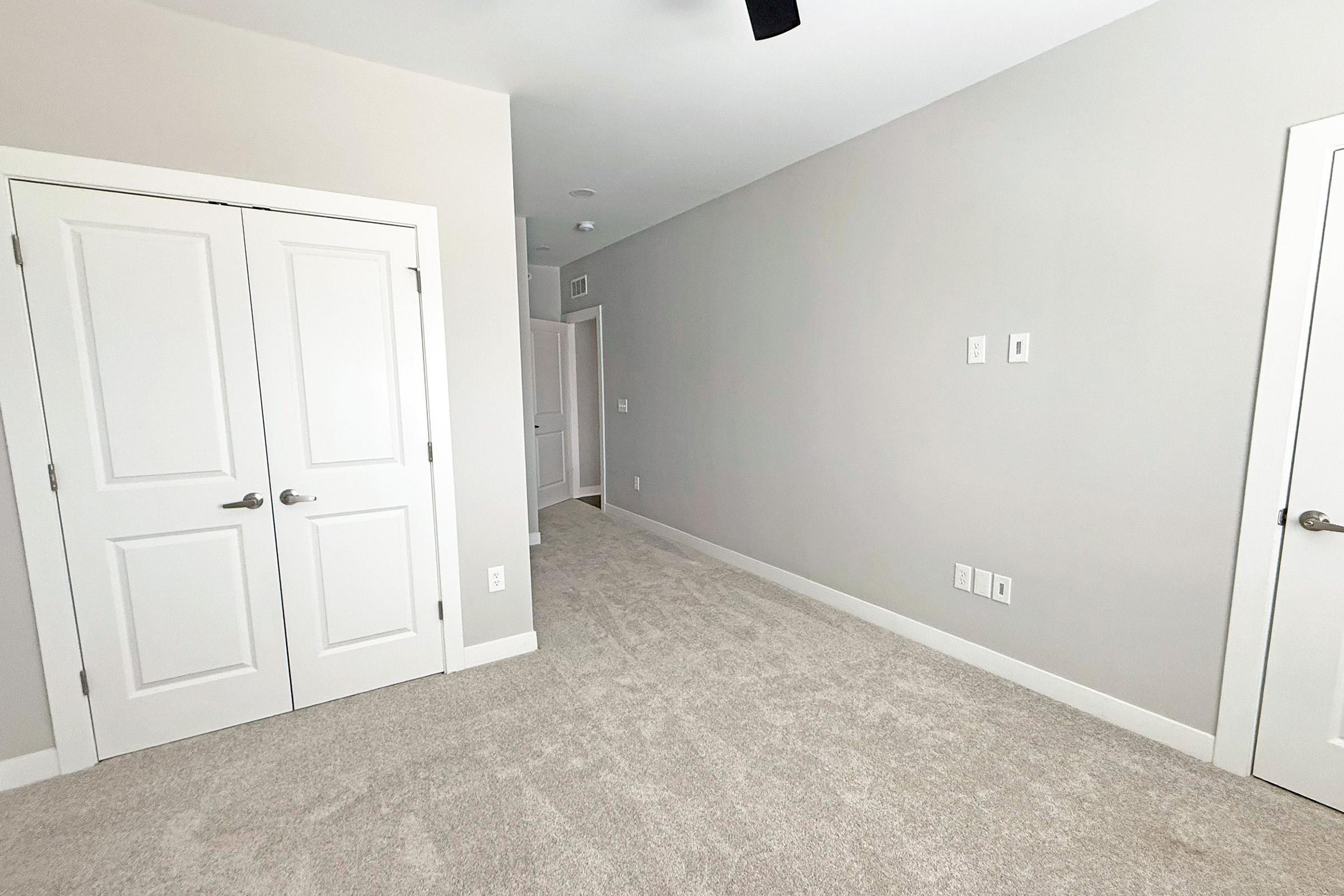 A light gray room featuring two white doors on the left, with a carpeted floor. The walls are painted in a soft gray color, creating a bright and modern atmosphere. A ceiling fan is visible, and there are electrical outlets along the wall. The space appears empty and well-lit.