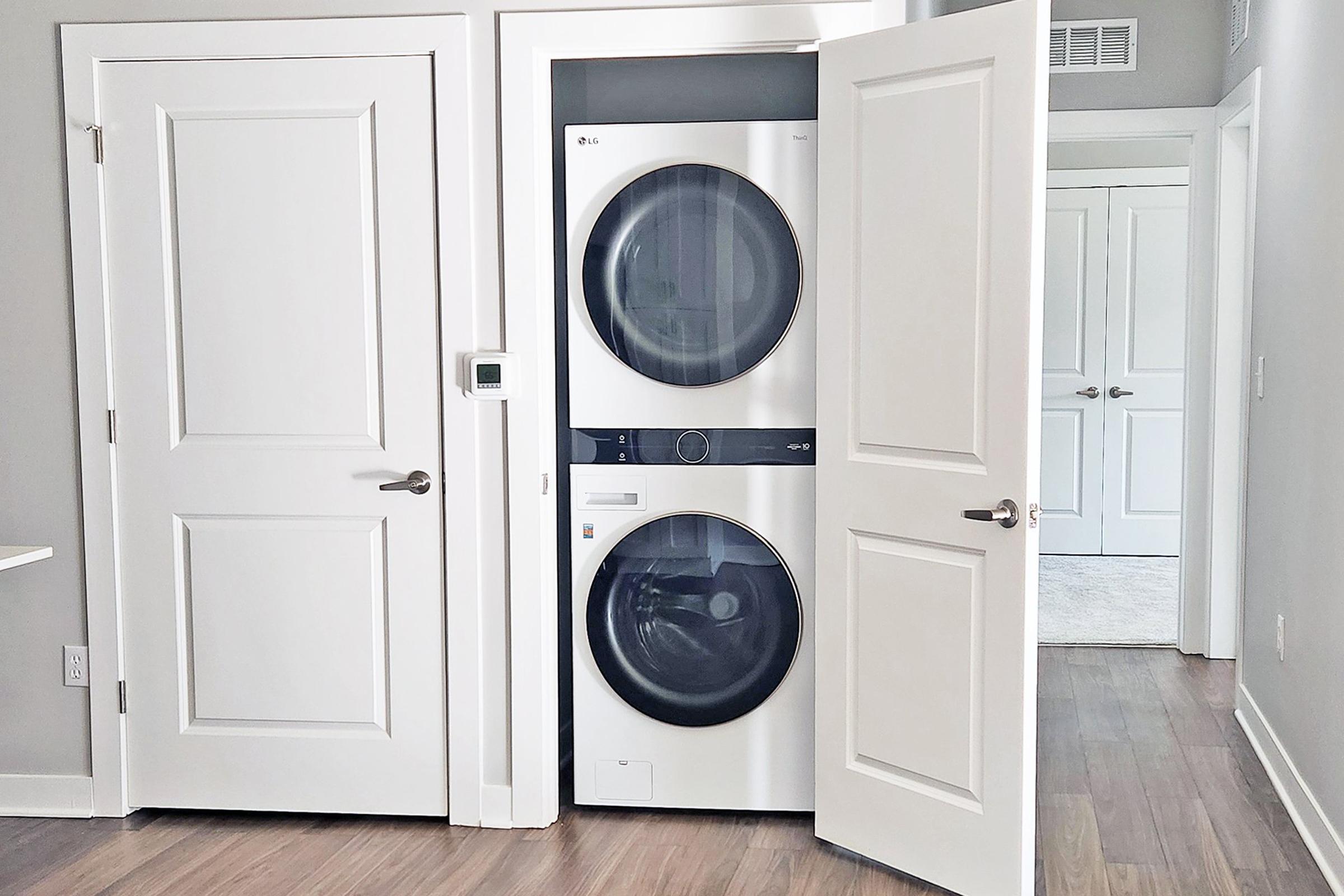 A modern laundry area featuring a stacked washer and dryer unit within a closet. The space has light gray walls and wooden flooring, with two white doors leading to adjacent rooms. A thermostat is visible on the wall beside the washer and dryer. The overall aesthetic is clean and contemporary.