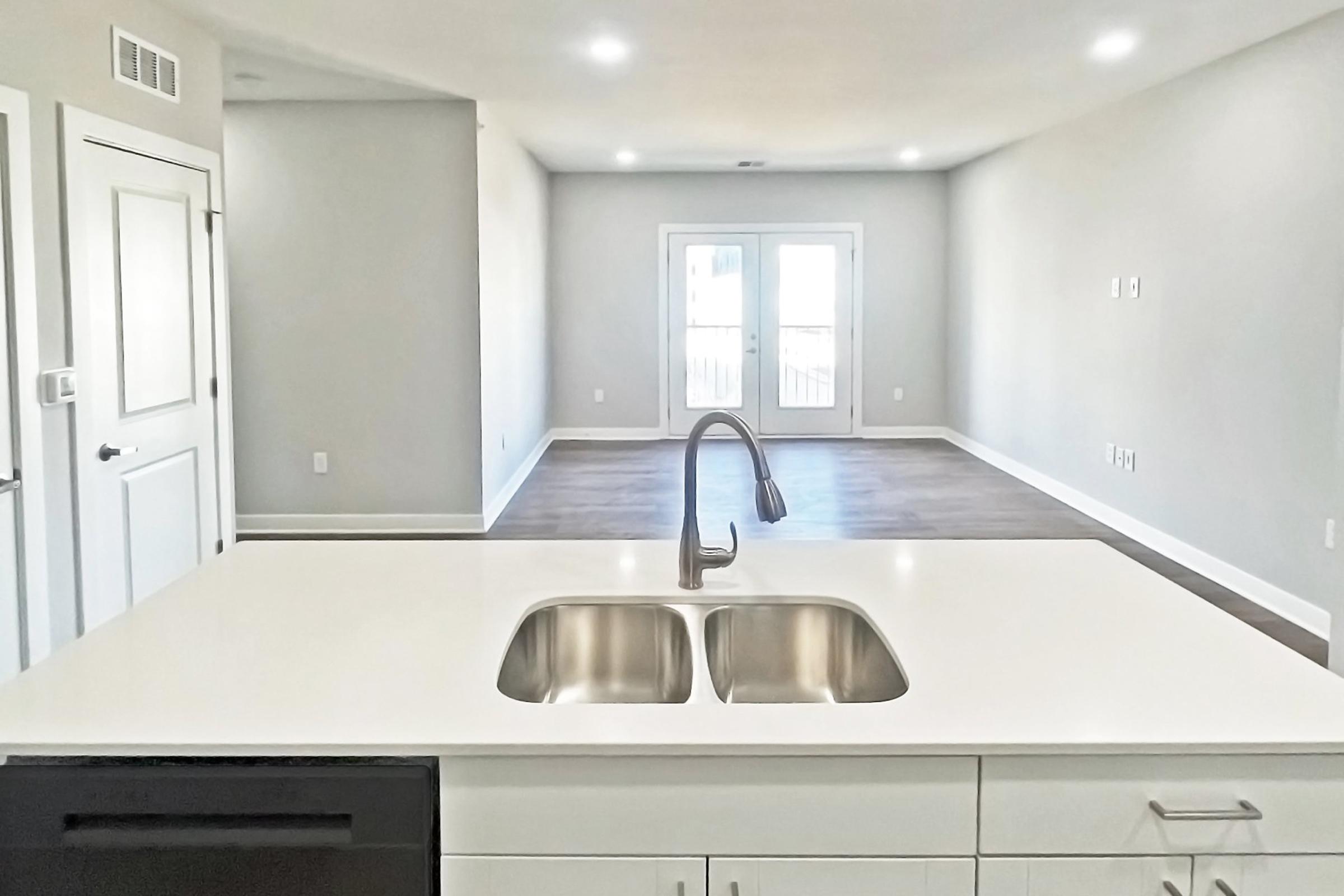 A modern kitchen with a white countertop and a double sink is in the foreground. The background shows a spacious, empty living room with light-colored walls and hardwood flooring, featuring sliding glass doors leading to an outdoor area. Bright overhead lights illuminate the space.