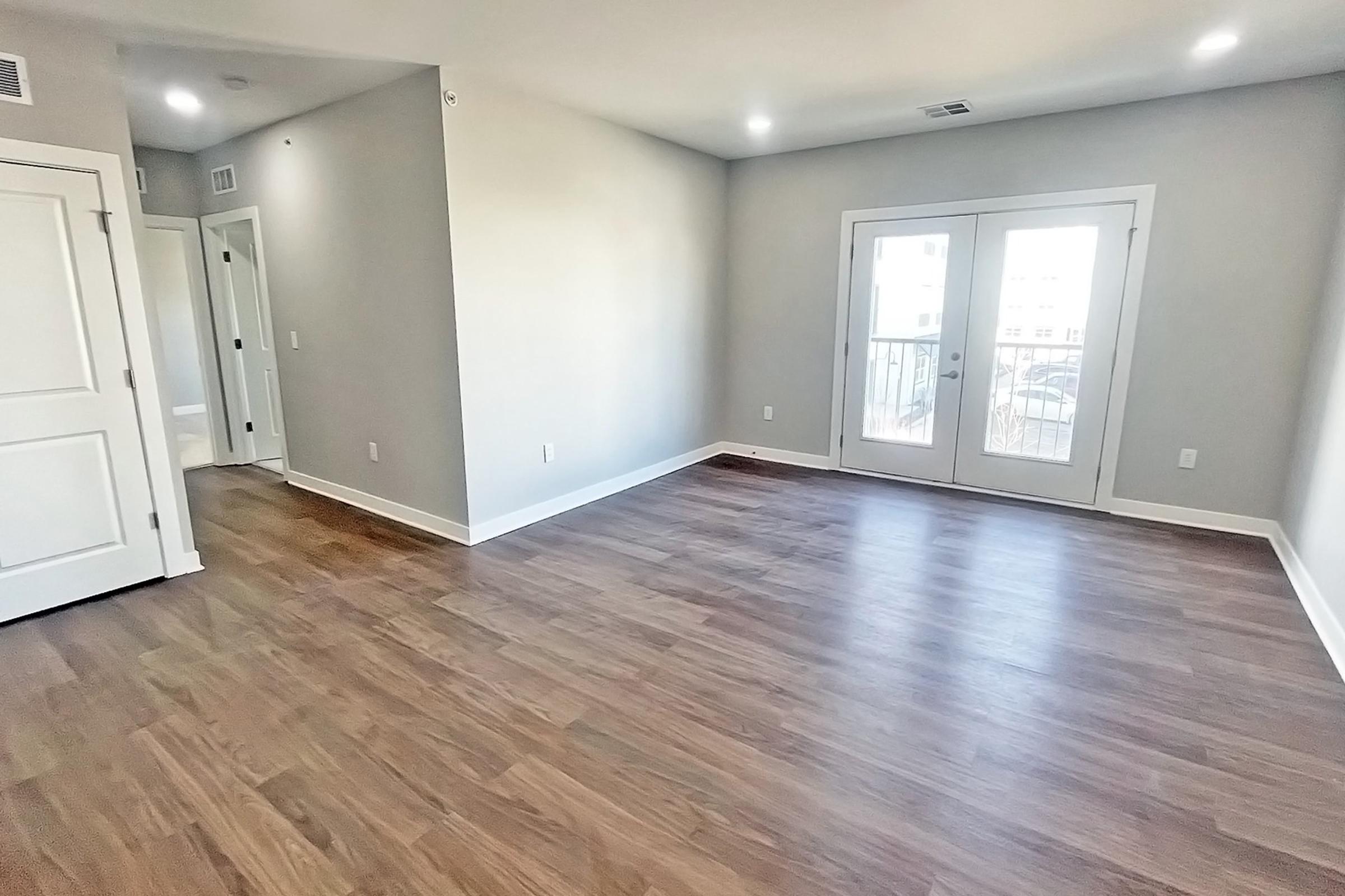 Empty modern living room with light-colored walls and wooden flooring. Large windows with French doors lead to a balcony, allowing natural light to fill the space. A hallway on the left leads to other rooms, and there are minimal furnishings, creating an open and airy atmosphere.