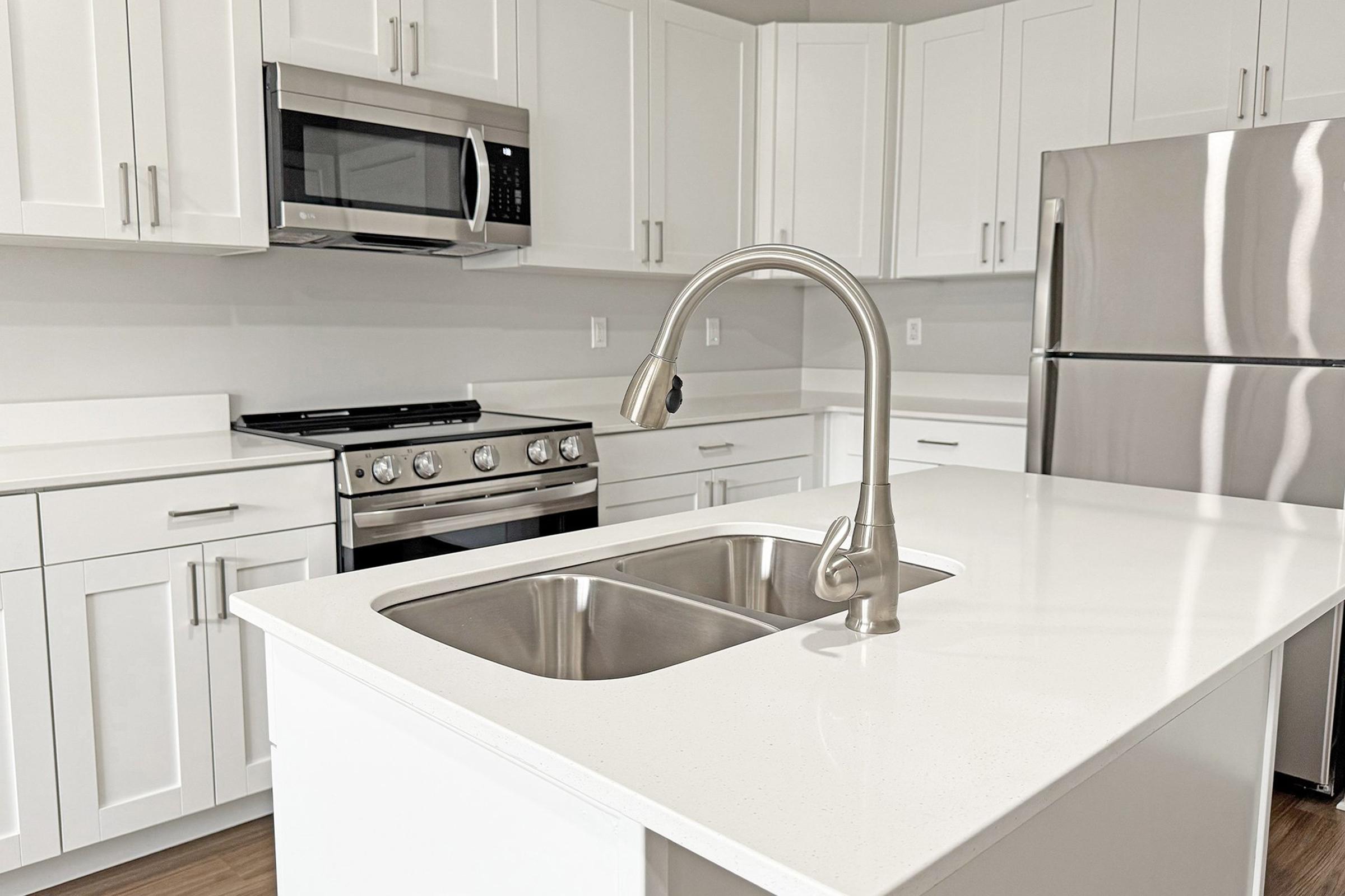 Modern kitchen featuring white cabinetry, a stainless steel sink with a sleek faucet, a gas stove, a microwave built into the cabinetry, and a stainless steel refrigerator. The countertop is light-colored quartz, and the overall aesthetic is clean and contemporary.