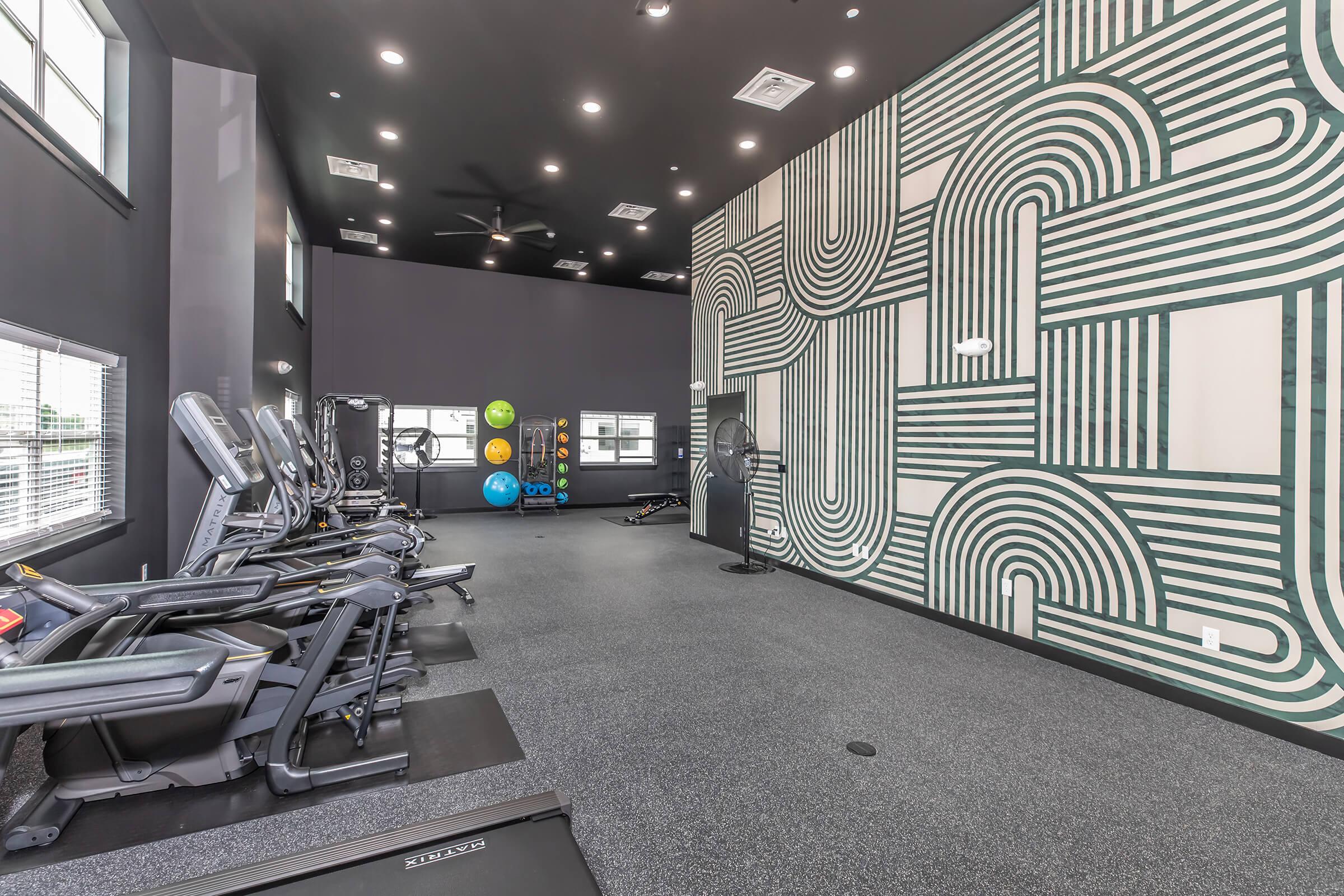 A modern gym interior featuring fitness equipment like treadmills and stationary bikes. The walls are adorned with a geometric mural in various shades, creating a vibrant atmosphere. Large windows allow natural light to fill the space, enhancing the energetic vibe of the workout area.