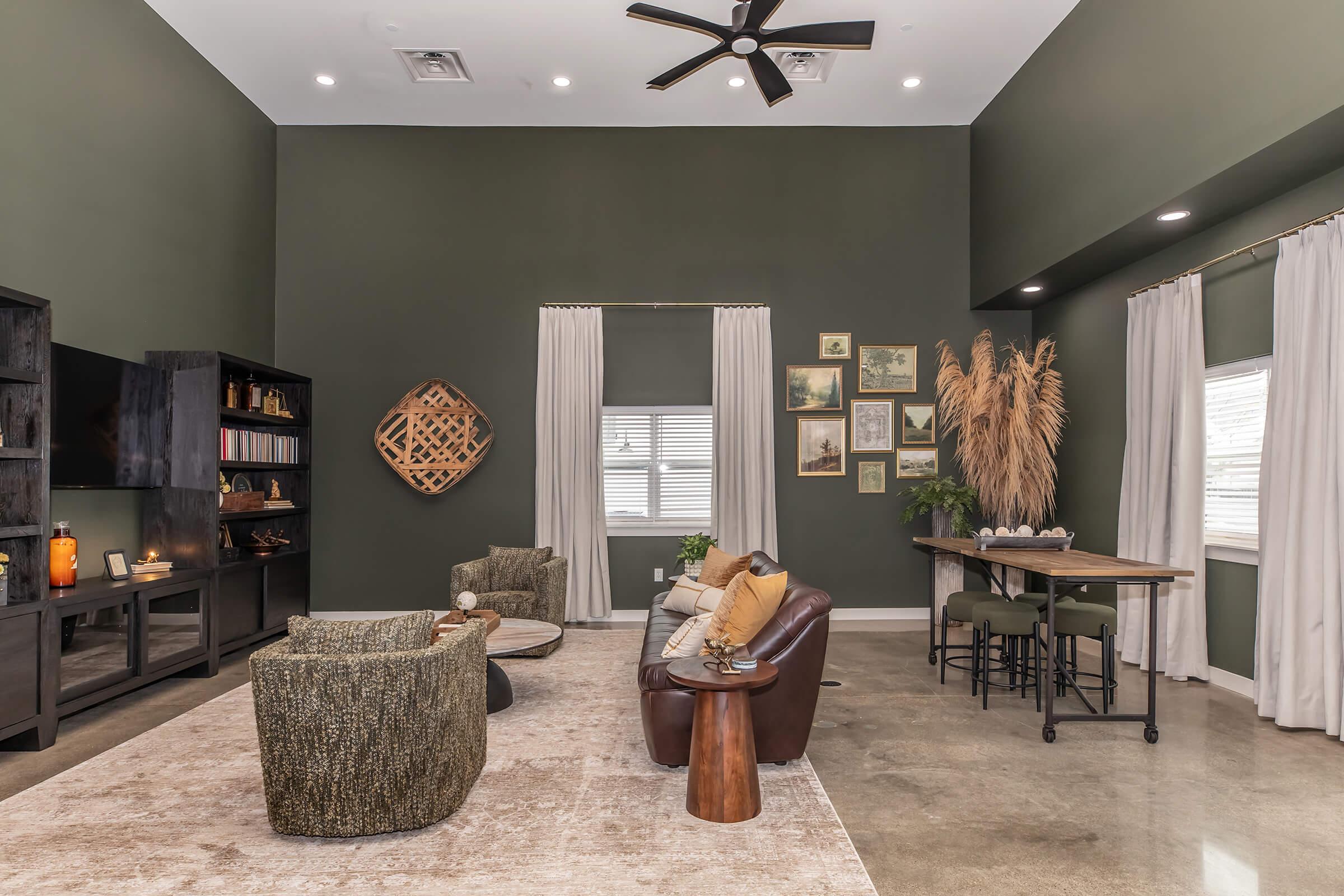A modern living room featuring dark green walls, a cozy seating area with a brown leather sofa and textured chairs, a wooden coffee table, and a sleek black bookshelf. Natural light streams in through large windows, complemented by decorative plants and framed artwork on the walls.