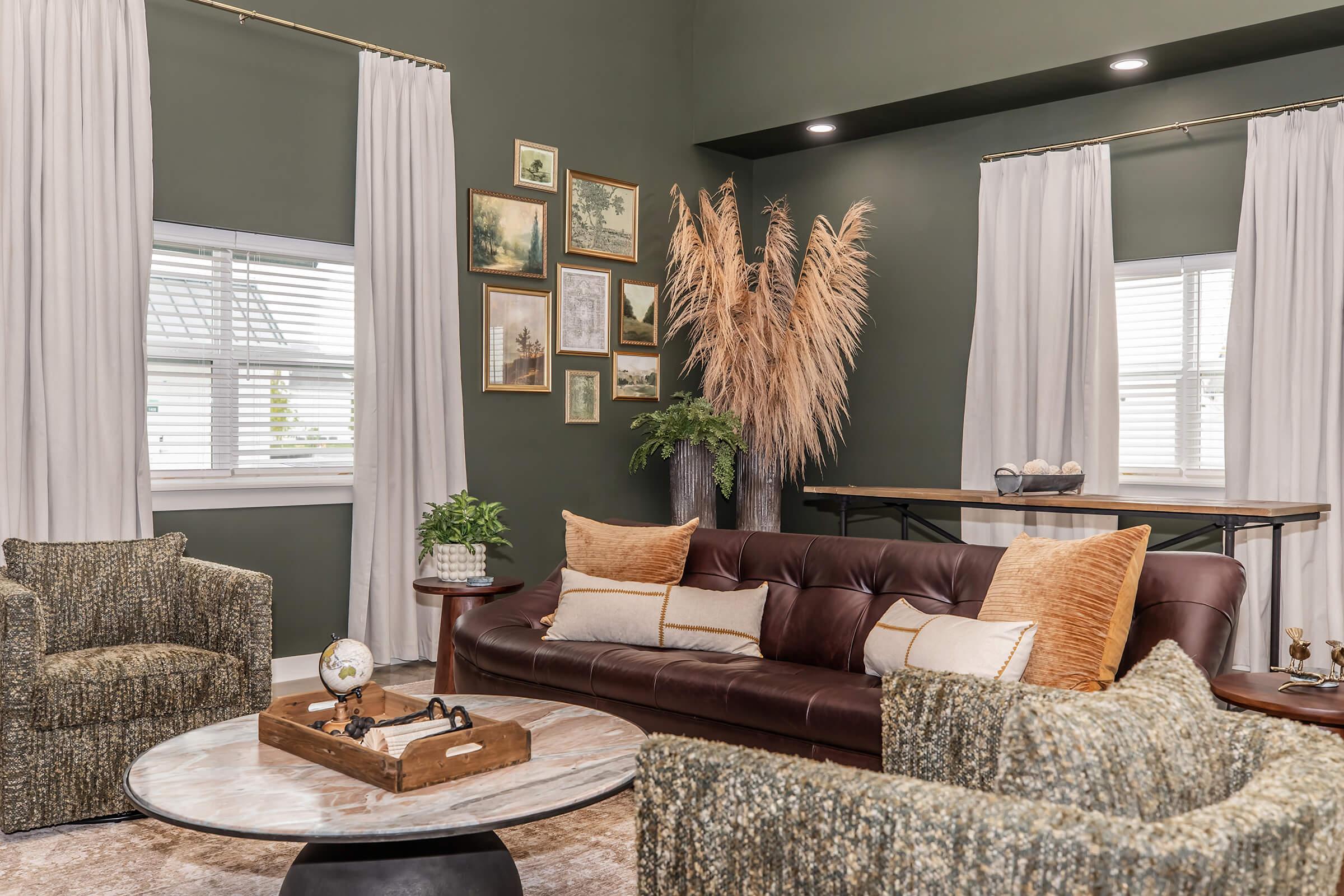 A modern living room featuring a dark green wall, large windows dressed with white curtains, and a cozy brown sofa with decorative pillows. Two textured armchairs are on either side of a round coffee table. A large pampas grass arrangement adds a natural touch, complemented by framed art on the walls and a decorative globe on the table.