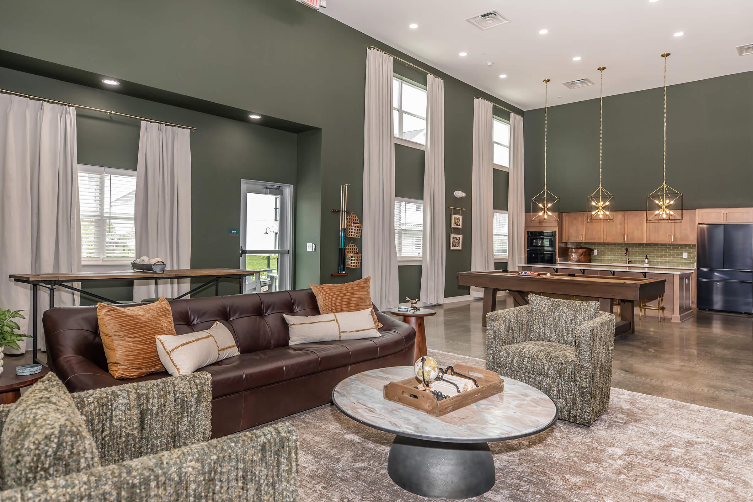 A stylish living space featuring a brown leather sofa with decorative pillows, two textured armchairs, and a round marble-top coffee table. The room has large windows with light curtains, a modern kitchen with wooden cabinets, and pendant lighting, all set against green walls and polished concrete floors.