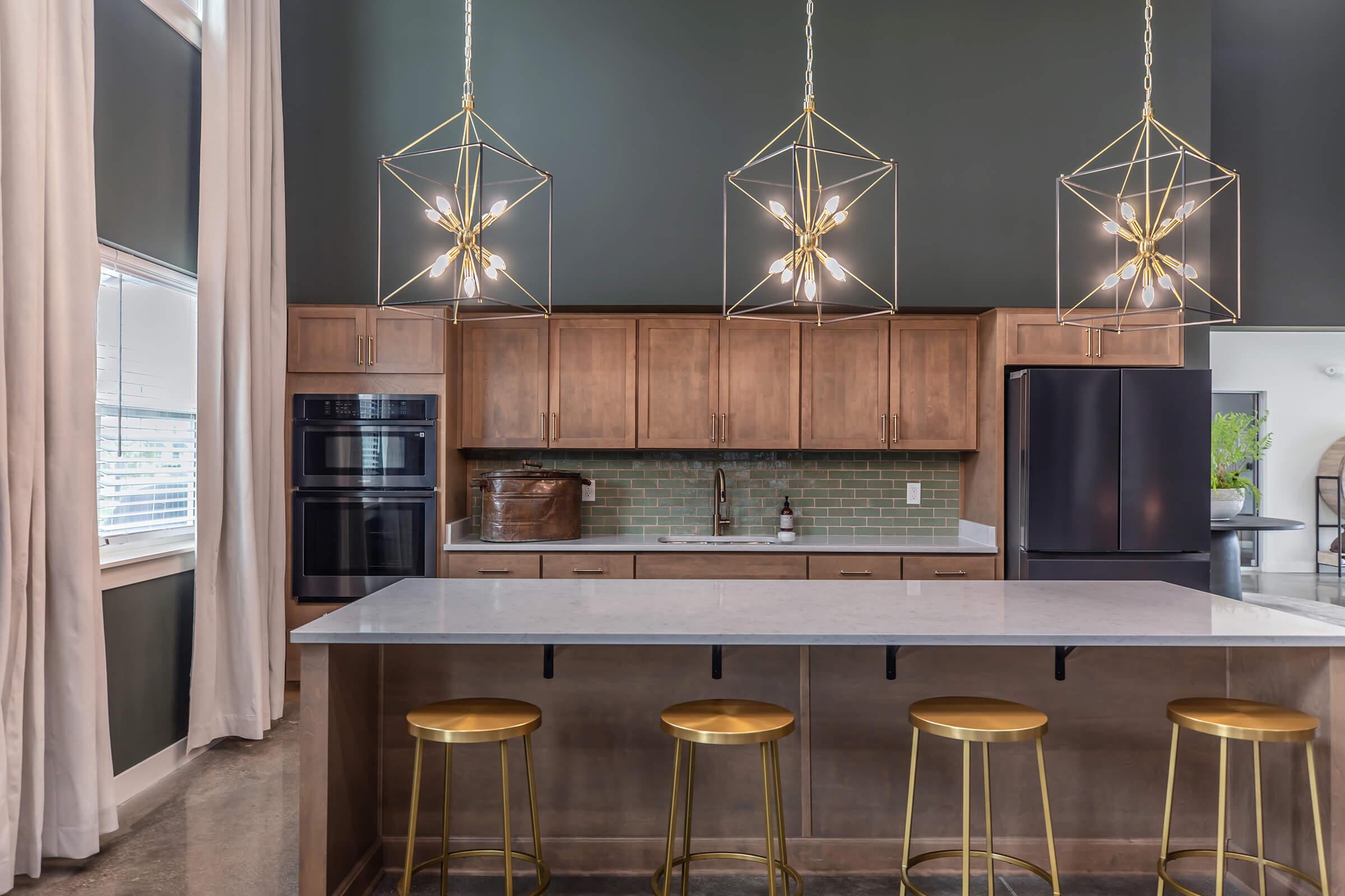 Modern kitchen featuring wooden cabinetry, stainless steel appliances, and a sleek island with four gold bar stools. The space is illuminated by geometric pendant lights and has large windows for natural light, complemented by a dark green accent wall and neutral-tone decor.