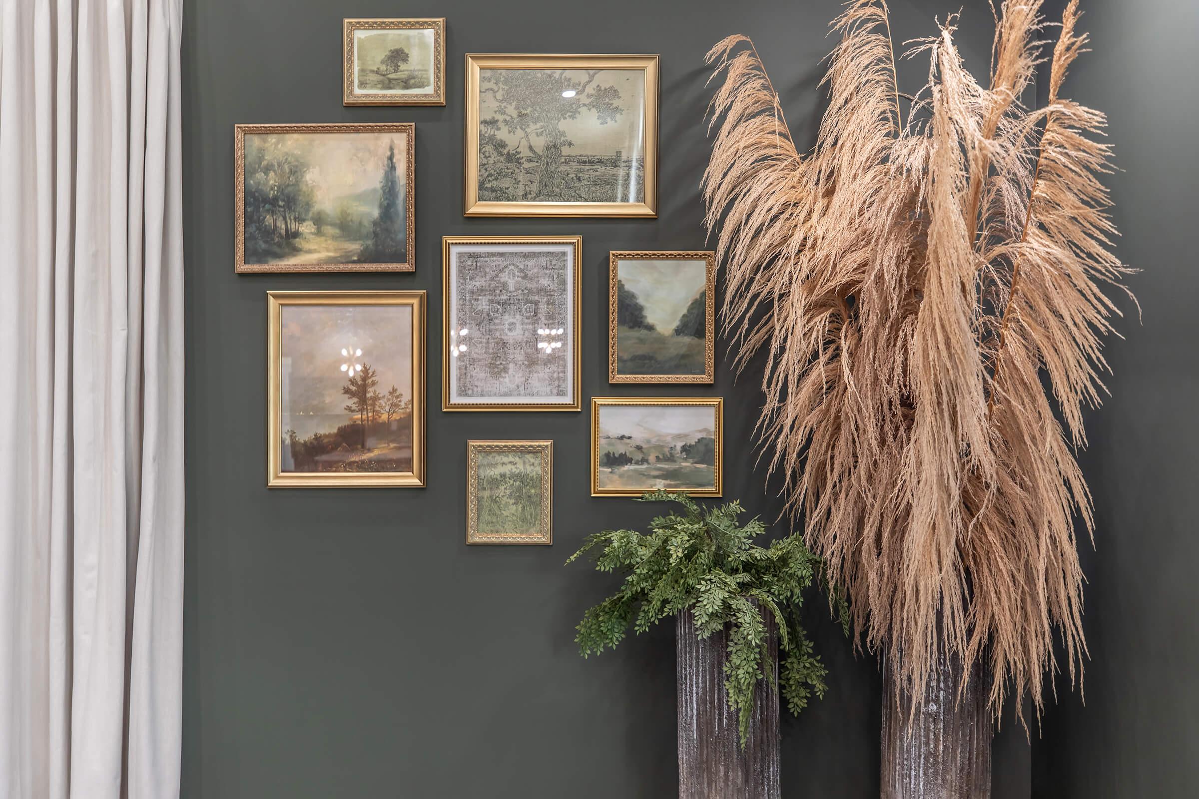 A modern interior wall featuring an arrangement of vintage-style framed paintings in gold frames. Adjacent to the artwork, there are two tall, textured planters containing dried pampas grass and lush green foliage, creating a harmonious blend of art and nature. Soft curtain drapes complement the decor.