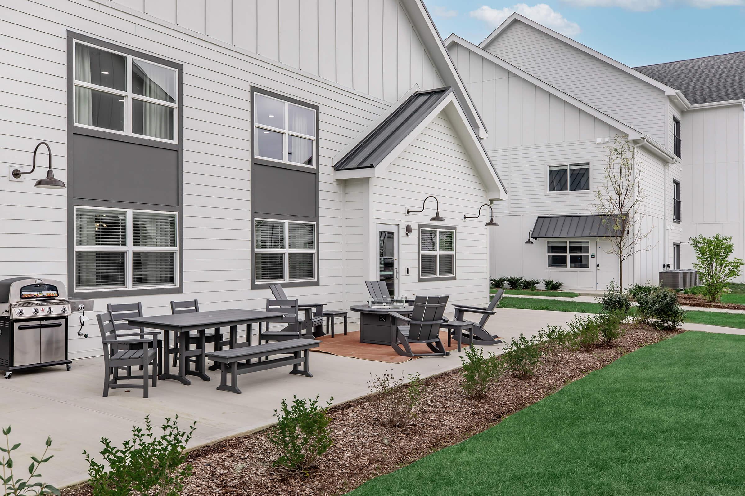 A modern outdoor patio area featuring a dining table with chairs, a grill, and a lounge space, set against a backdrop of a white two-story building with large windows. The landscape includes freshly cut grass and neatly arranged shrubs, creating an inviting atmosphere for gatherings and relaxation.