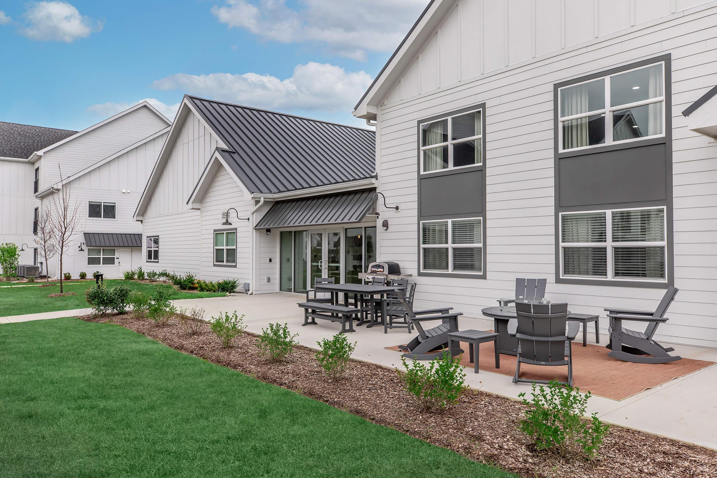 Exterior view of a modern residential building featuring a well-maintained lawn, a patio area with outdoor seating, and a barbecue grill. The building has a contemporary design with light-colored siding and a dark metal roof. Clear blue sky above enhances the inviting atmosphere.