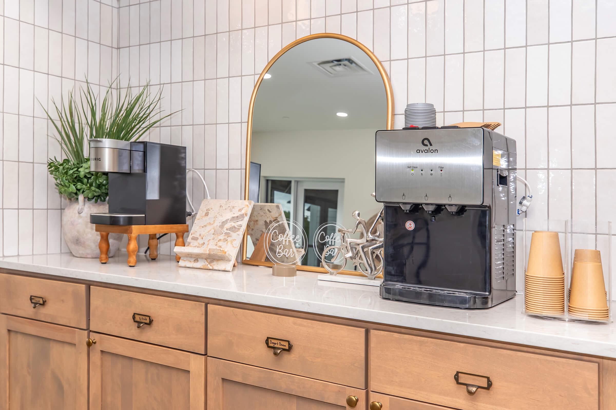 A stylish coffee station featuring a sleek black coffee machine, a decorative mirror, a potted plant, and a small stack of coffee cups. The setup is on a counter with light-colored cabinetry, showcasing modern design elements and a cozy ambiance.