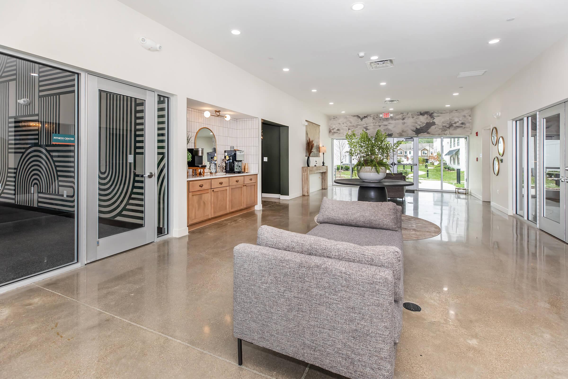 Welcoming lobby area featuring modern decor. A gray sofa is positioned in the foreground, while the walls have a minimalist design. To the side, there is a coffee station with countertop and mirrors. Large windows provide natural light, opening to an outdoor view, enhancing the spacious and inviting atmosphere.