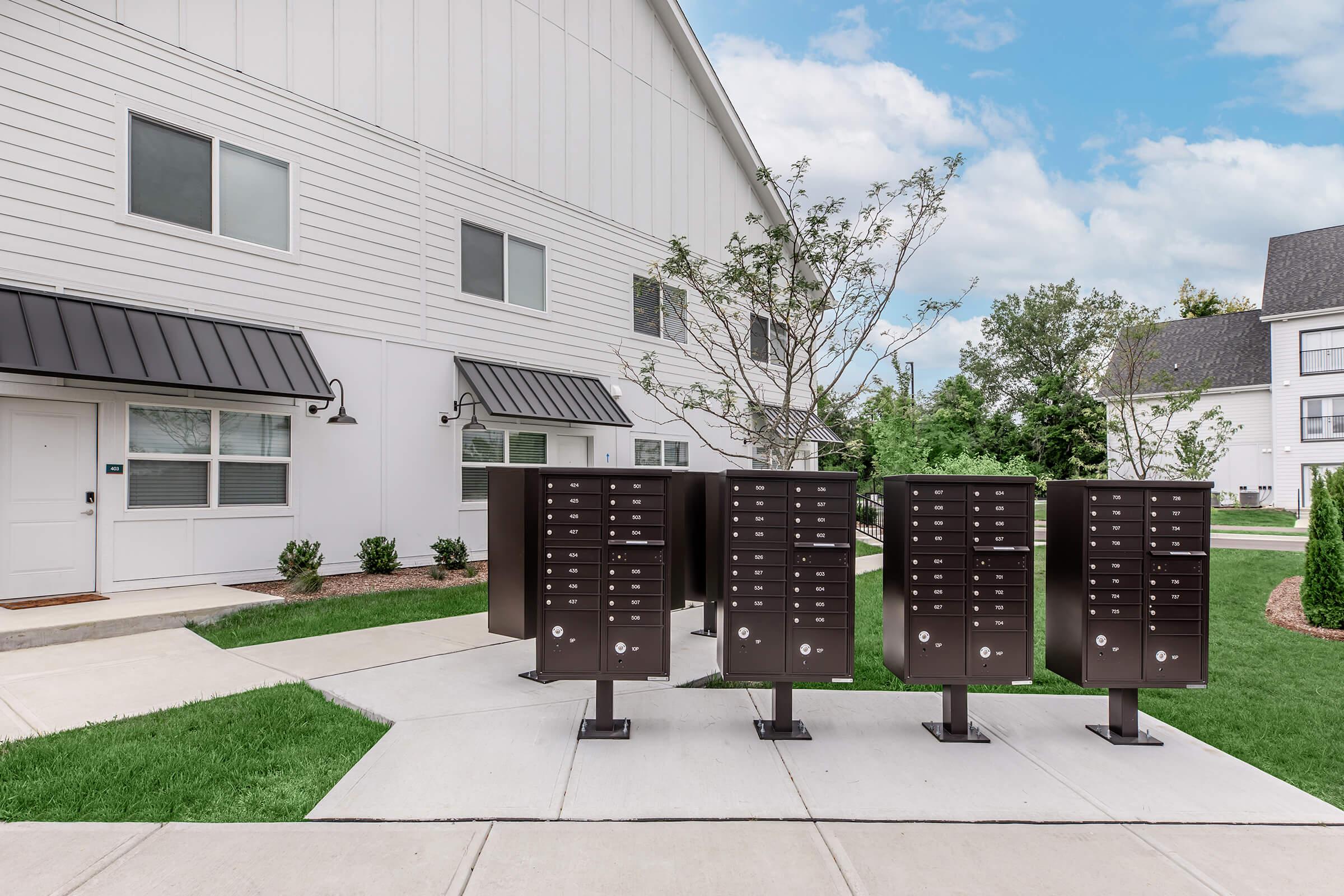 A cluster of black outdoor mailboxes standing on a paved walkway in front of a modern residential building. Green grass and small shrubs are present around the area, with a clear blue sky in the background. The building features a white facade with dark roofing and large windows.