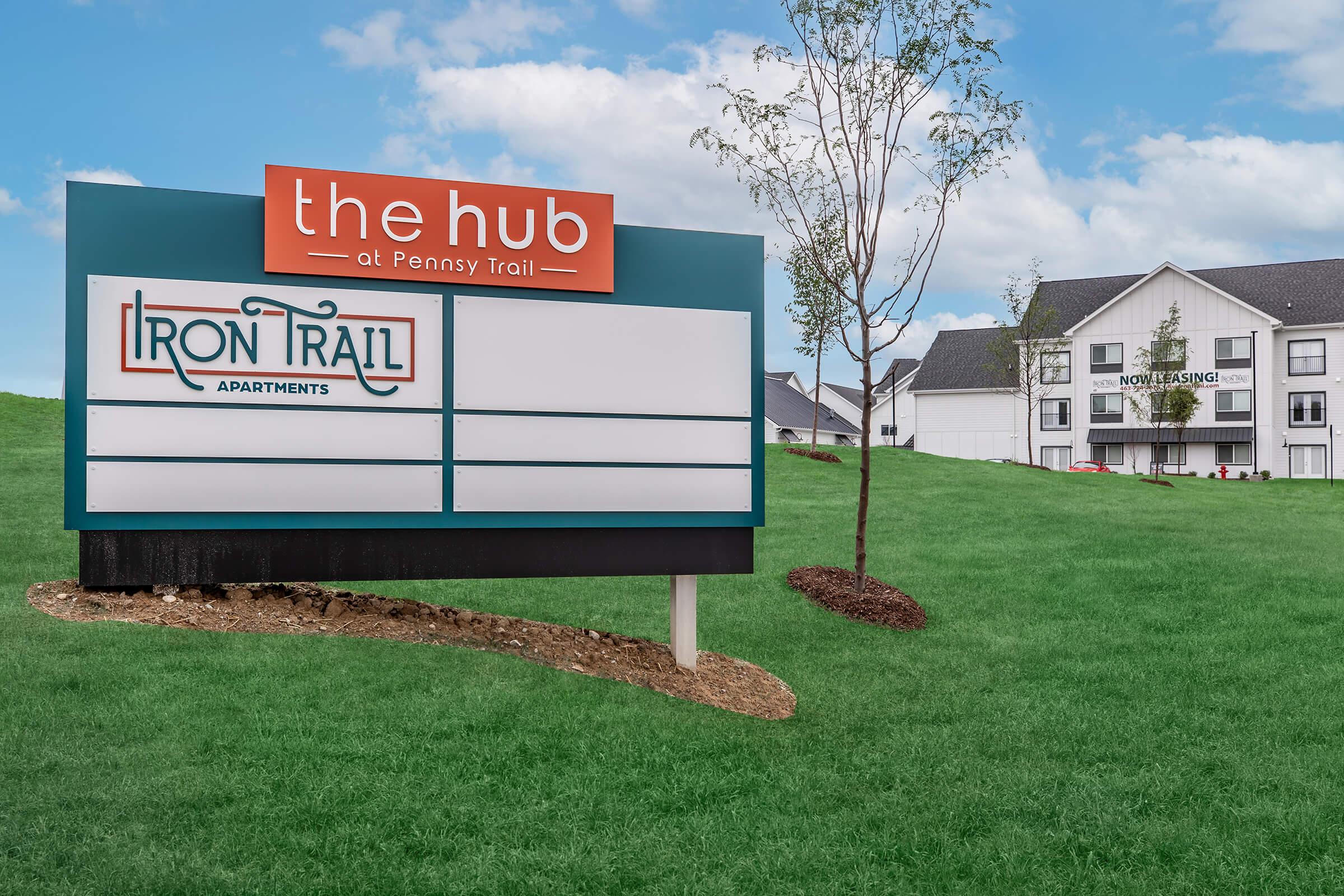 Sign for "the hub at Penny Trail" with "IRON TRAIL APARTMENTS" listed below. The sign is in front of a grassy area with trees, and an apartment building is visible in the background. The sky is clear with soft clouds.
