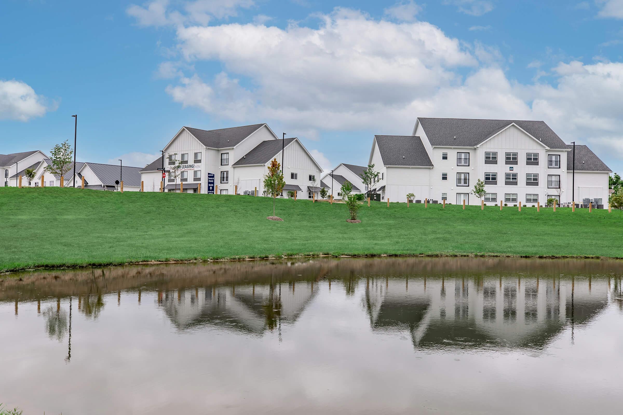 A serene landscape featuring modern white homes with dark roofs beside a calm pond. The reflection of the houses is visible in the water, surrounded by lush green grass and a partly cloudy blue sky. The scene conveys a tranquil suburban atmosphere.