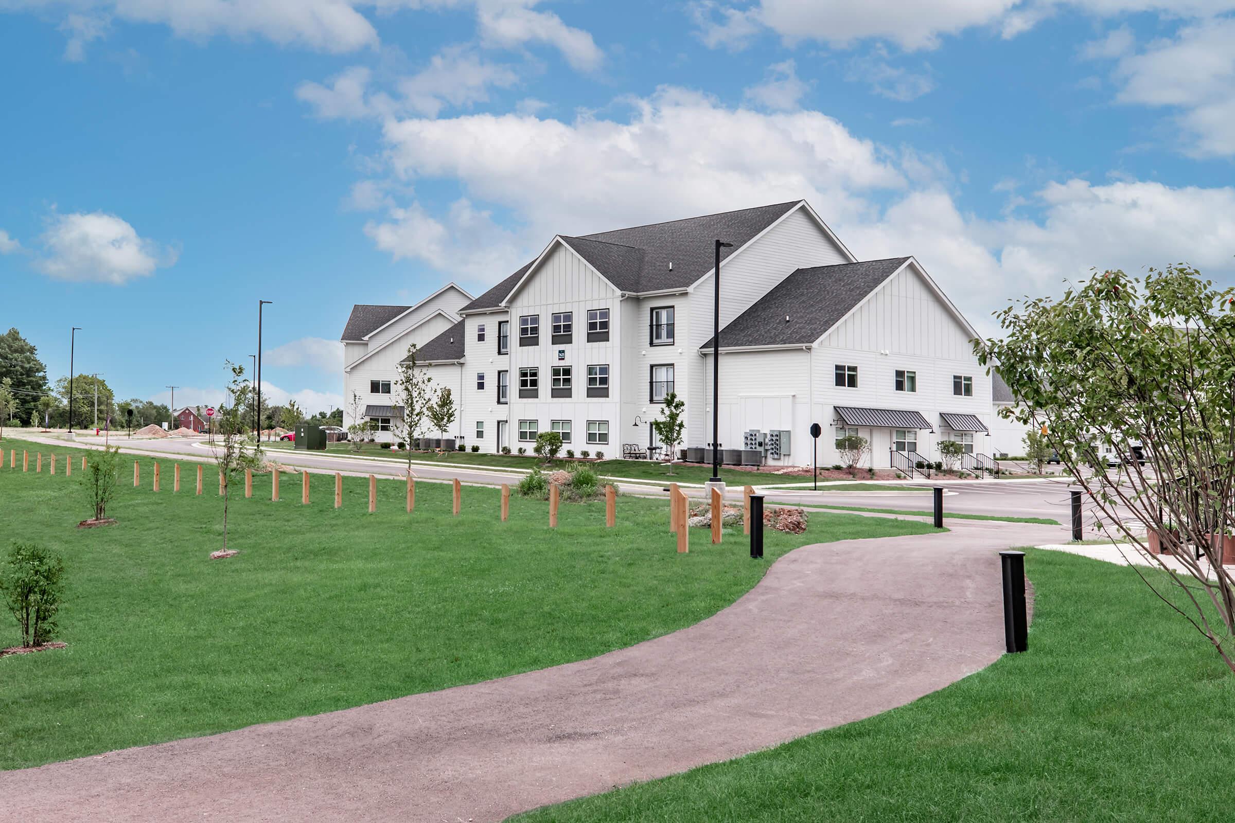 A modern multi-story building with a white exterior is set amidst a landscaped area featuring green lawns, small trees, and a walking path. The sky is blue with fluffy clouds, creating a serene environment. The architecture includes large windows and a mix of residential and community elements.
