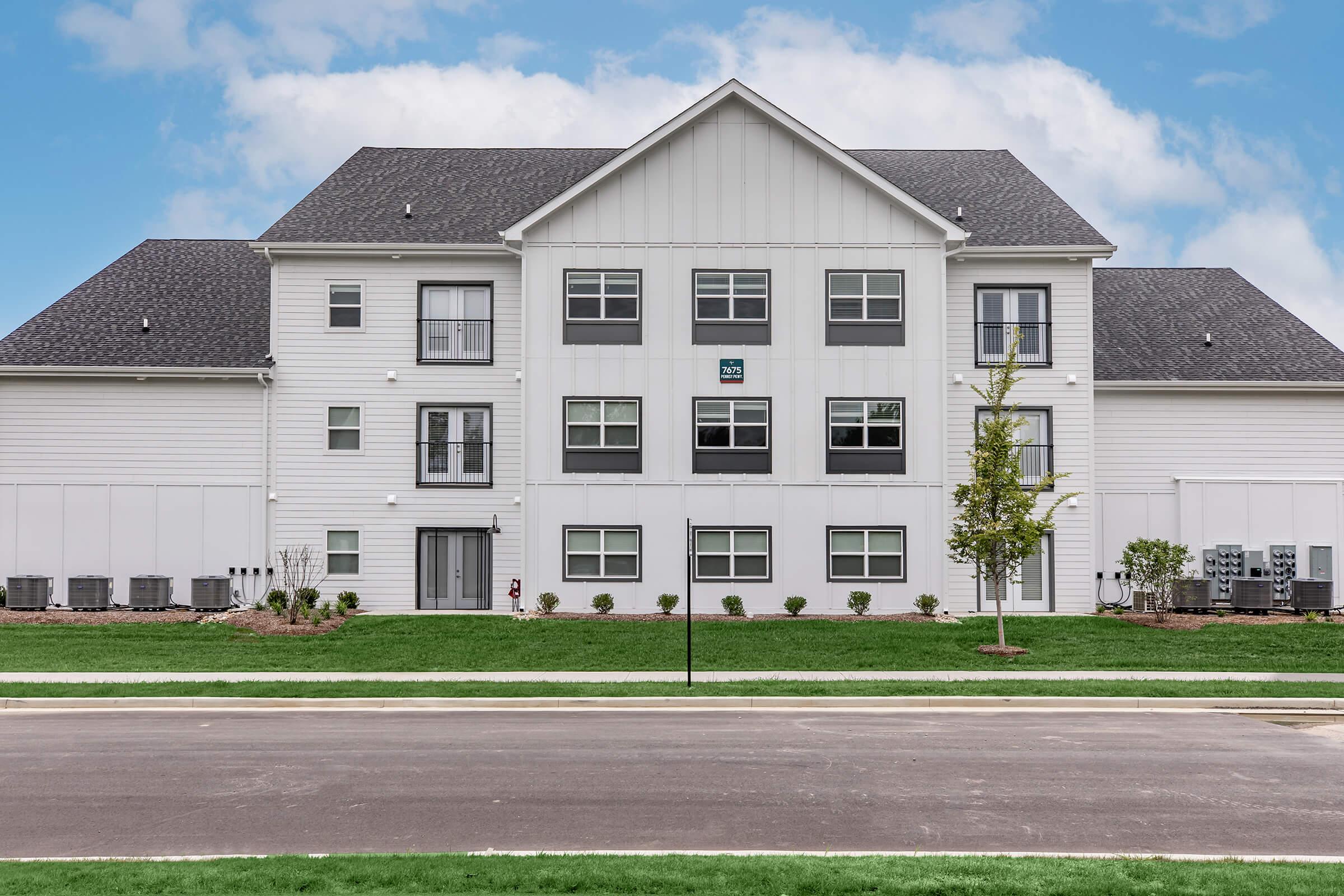 A modern two-story residential building with a light-colored exterior and dark roof, featuring several windows on the front. The building is surrounded by green grass, small bushes, and trees. In front, there is a paved street with no visible vehicles. The sky is clear with a few clouds.