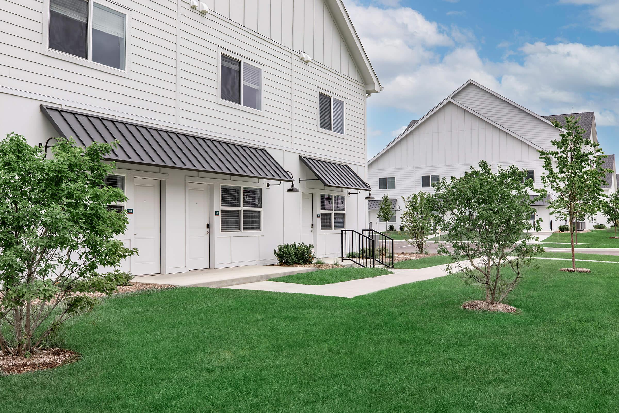 A modern residential building exterior featuring two two-story structures with white siding, black metal awnings over doors, and landscaped walkways. The foreground includes grass and small trees, with a cloudy blue sky in the background.