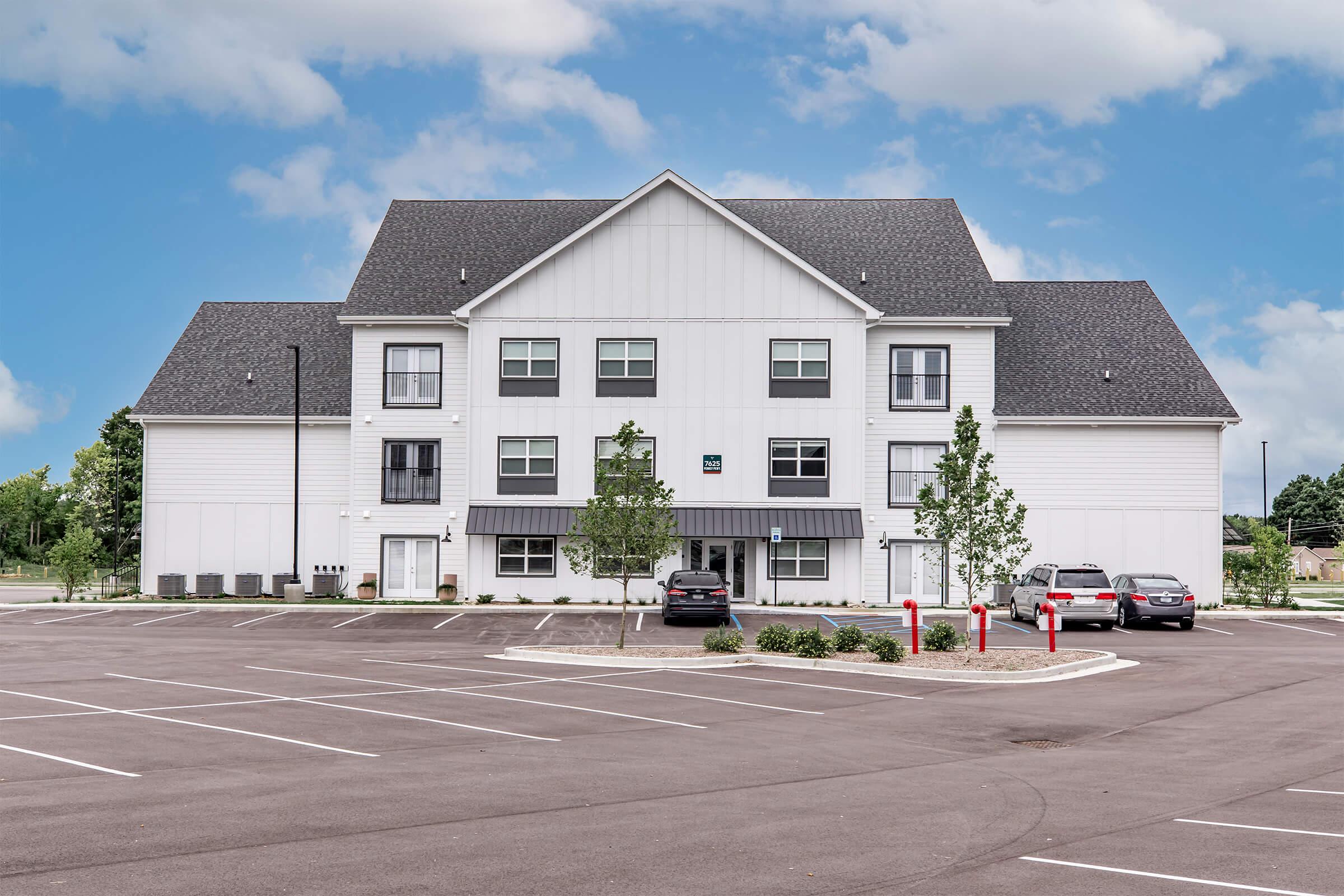 Modern three-story hotel building with a white facade and dark roofing. Surrounding the building are neatly trimmed trees and a spacious parking lot with several parked cars. The sky is partly cloudy, adding to the serene atmosphere of the location.