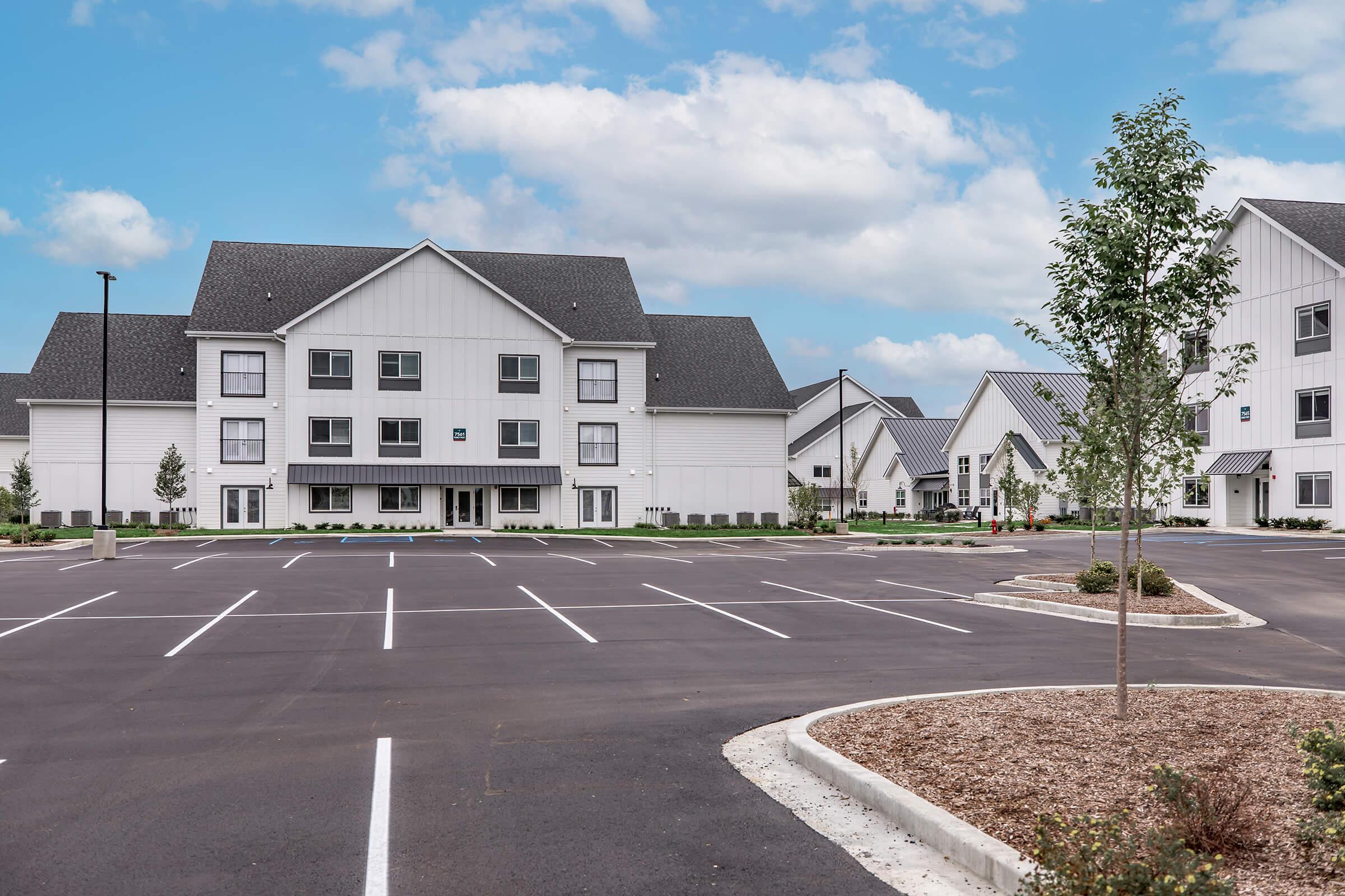 Modern white multi-story building with gray roofs, surrounded by a landscaped area and a large, empty parking lot. The sky is clear with a few clouds, and additional buildings can be seen in the background, creating a clean and inviting neighborhood atmosphere.