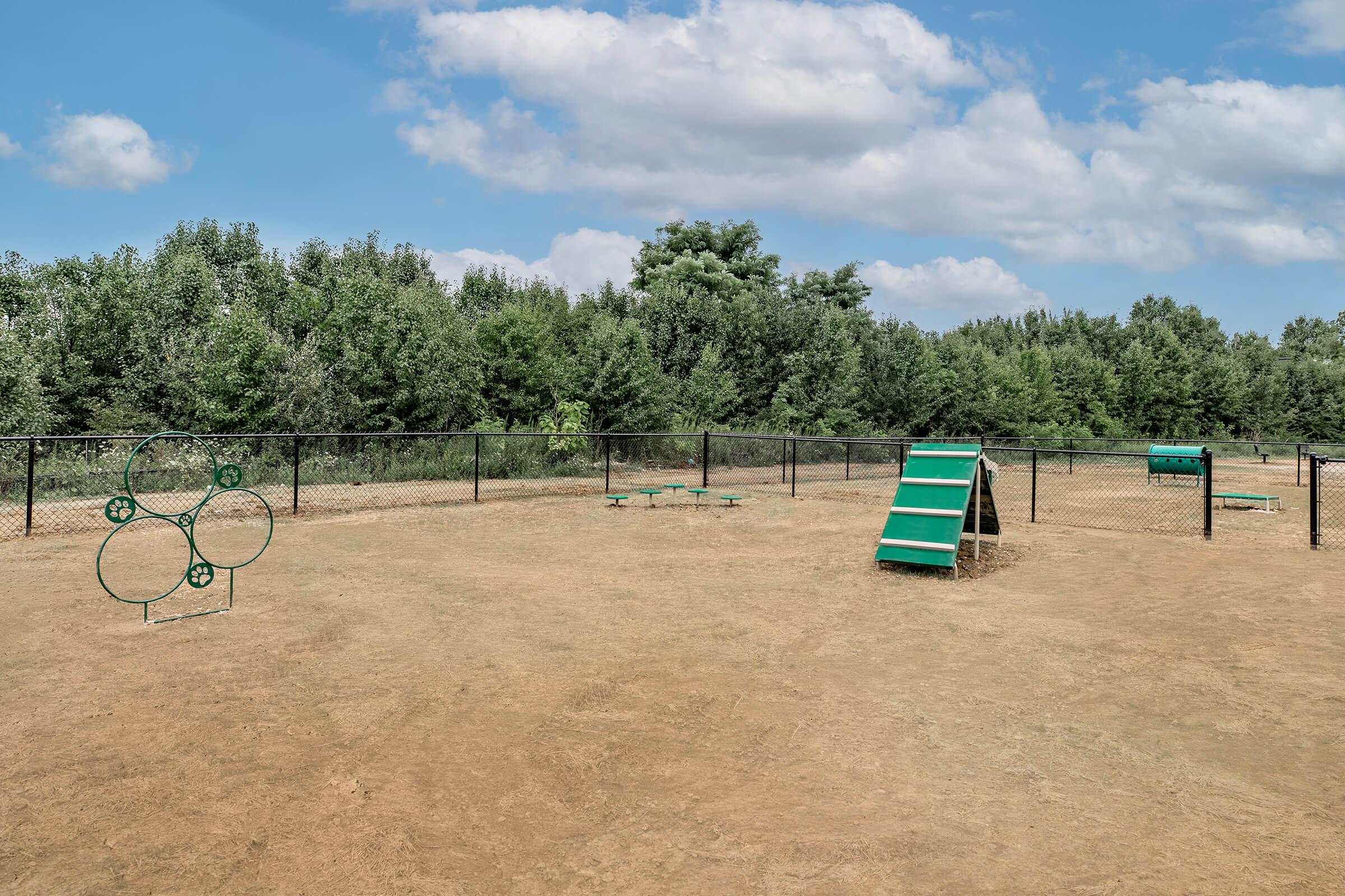 A dog park featuring a grassy area enclosed by a fence. In the foreground, there is a green agility tunnel and a rectangular climbing frame. A circular ring jump is visible nearby, with benches in the background and trees lining the area under a partially cloudy sky.