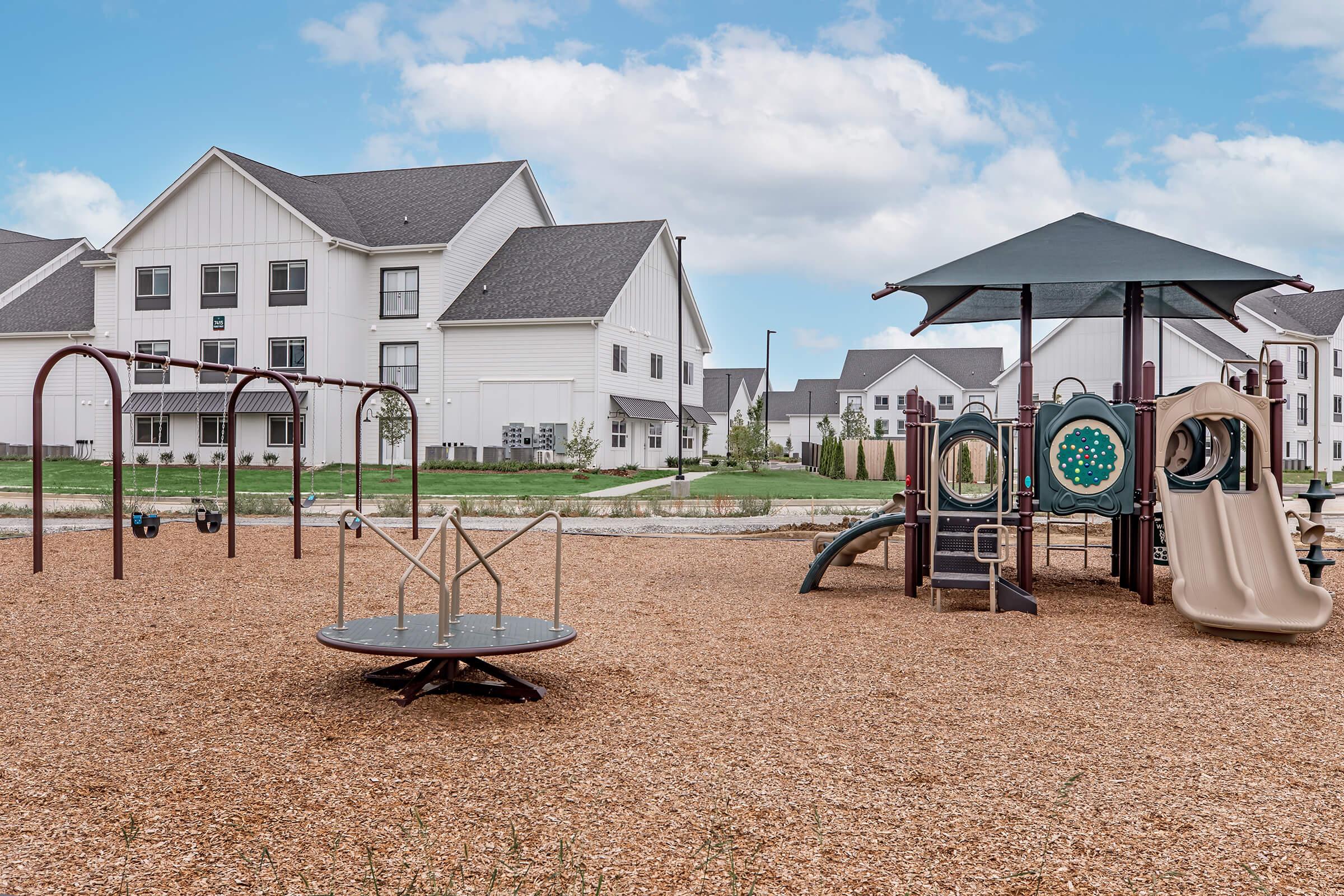 A playground featuring swings, a merry-go-round, and a slide, situated on a bed of wood chips. In the background, there are residential buildings with a clear blue sky overhead. The area appears inviting for children to play.