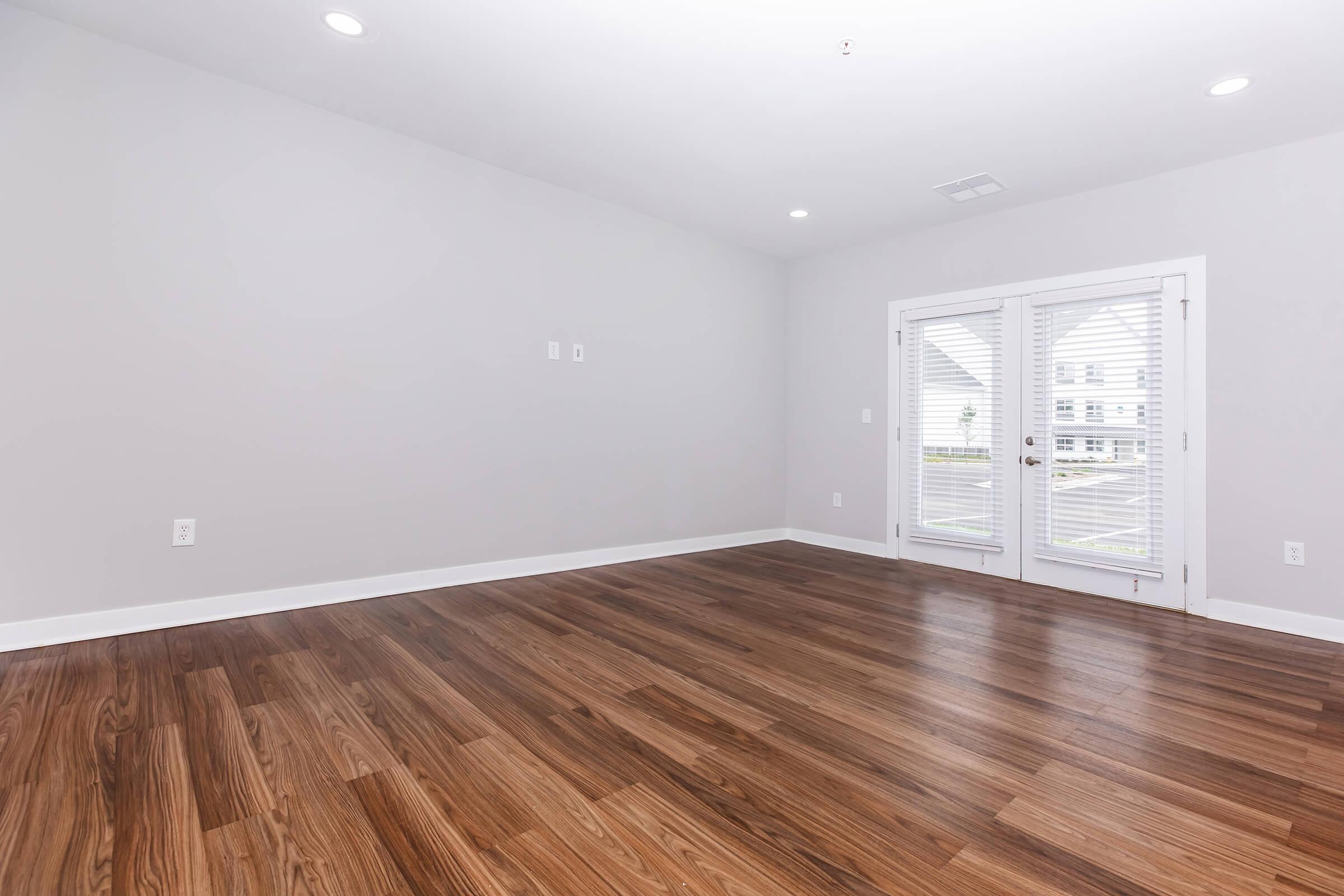 An empty room with light gray walls and a wooden floor. There are two glass French doors with white blinds leading to an outdoor area. The lighting is bright, highlighting the spaciousness of the room.