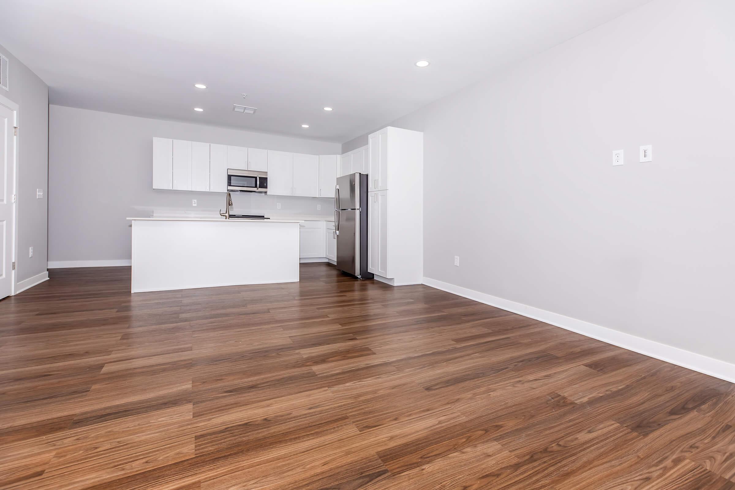 A modern, empty kitchen and living area with light-colored walls and hardwood flooring. The kitchen features white cabinets, an island, and stainless steel appliances, including a microwave and refrigerator. Recessed lighting is present, creating a bright and spacious ambiance.