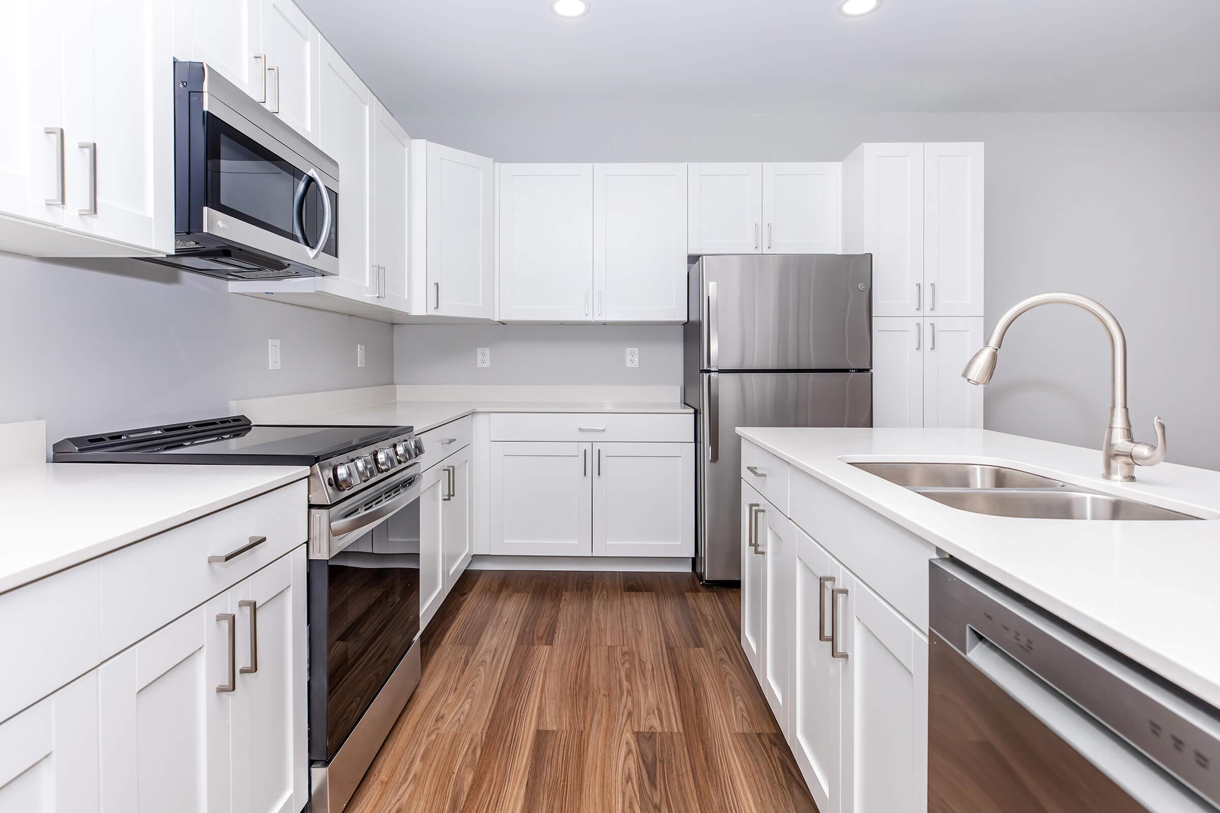 Modern kitchen featuring white cabinetry, stainless steel appliances including a microwave and refrigerator, a sleek gas stove, and a large sink. The space has a light and airy feel with hardwood flooring and bright overhead lighting.