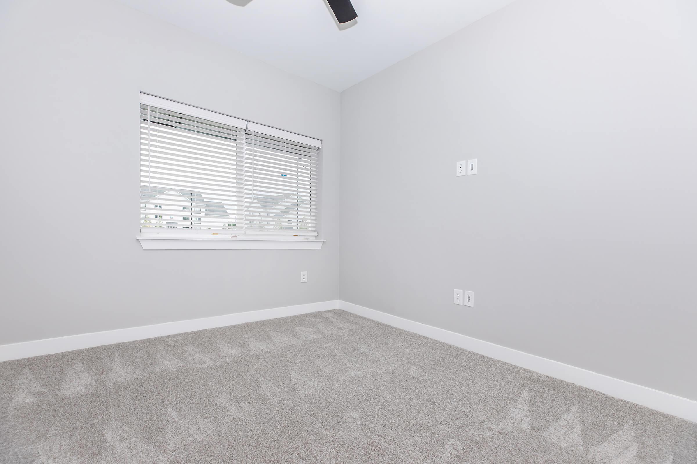 A vacant room with light gray walls and a ceiling fan. The floor is covered in soft carpet, and there is a window with white blinds allowing natural light to enter. The walls have two electrical outlets but no furniture or decorations in sight.