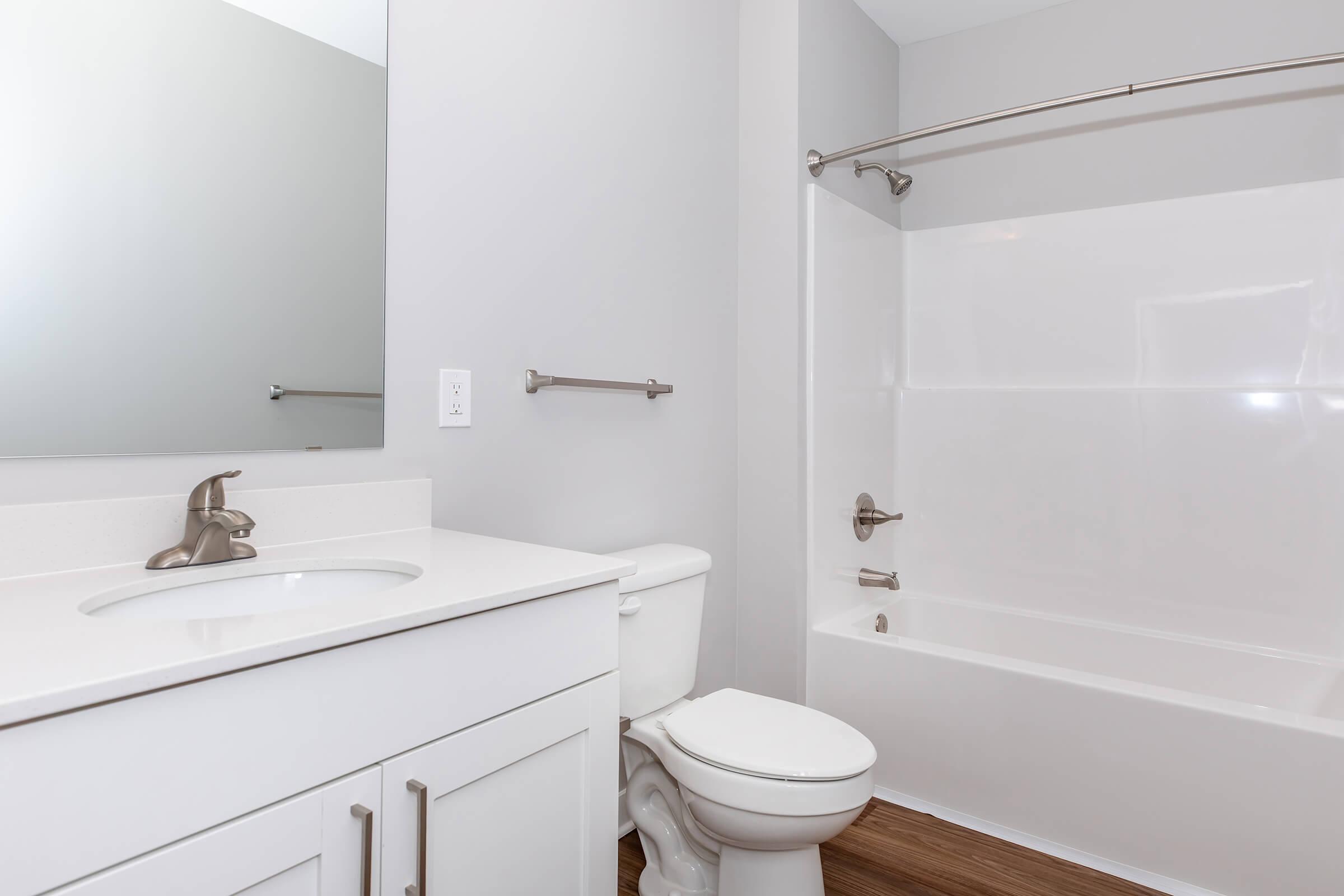 A modern bathroom featuring a white bathtub and shower, a white vanity with a sink, and a toilet. The walls are painted light gray, and there is a large mirror above the sink. The floor has a wooden appearance, adding warmth to the space.