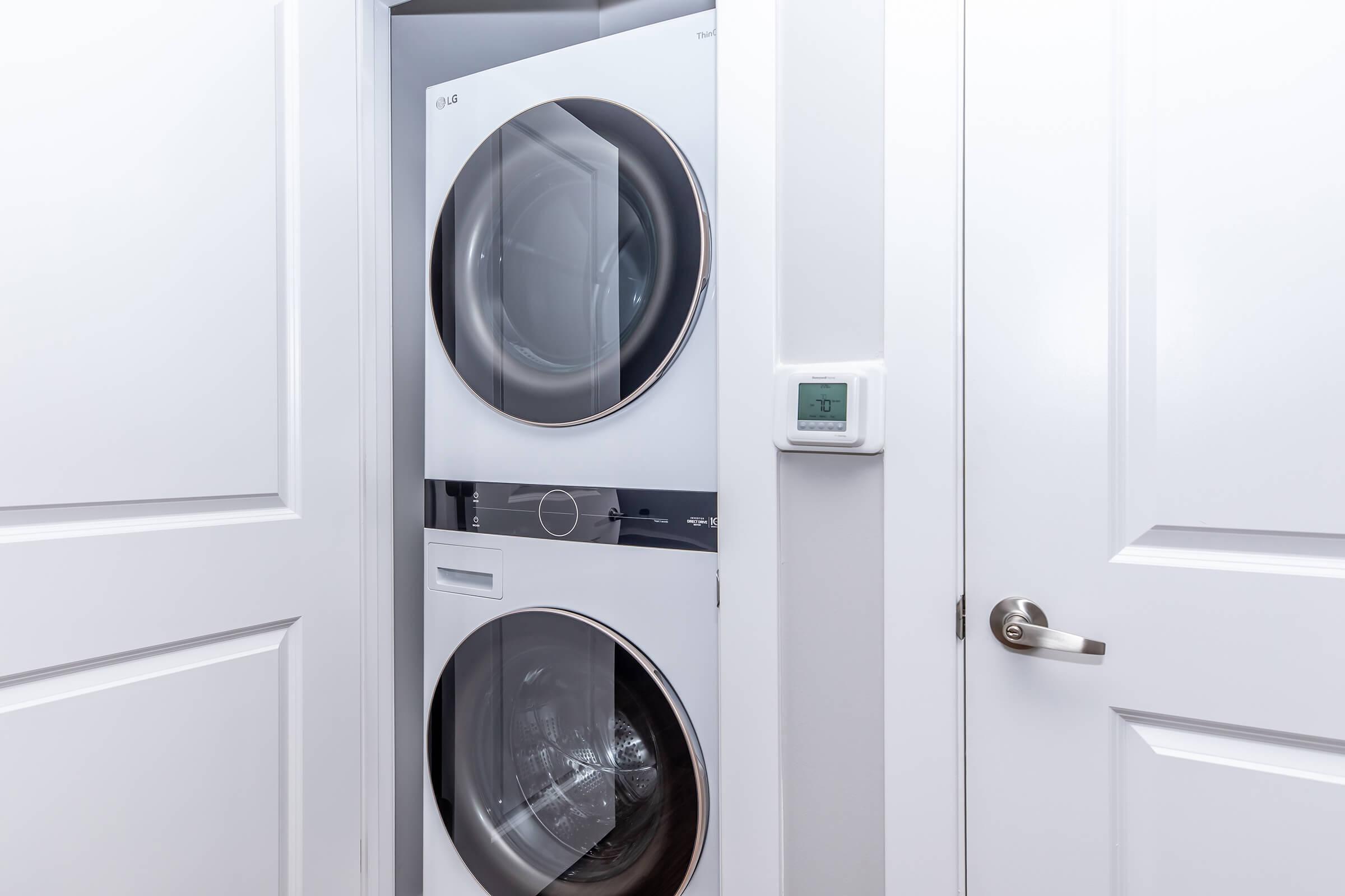 A stacked washer and dryer unit situated in a laundry closet, with a thermostat on the wall next to it. The doors of the closet are closed but partially ajar, giving a glimpse of the appliances inside. The overall setting is clean and modern, featuring white cabinetry and walls.