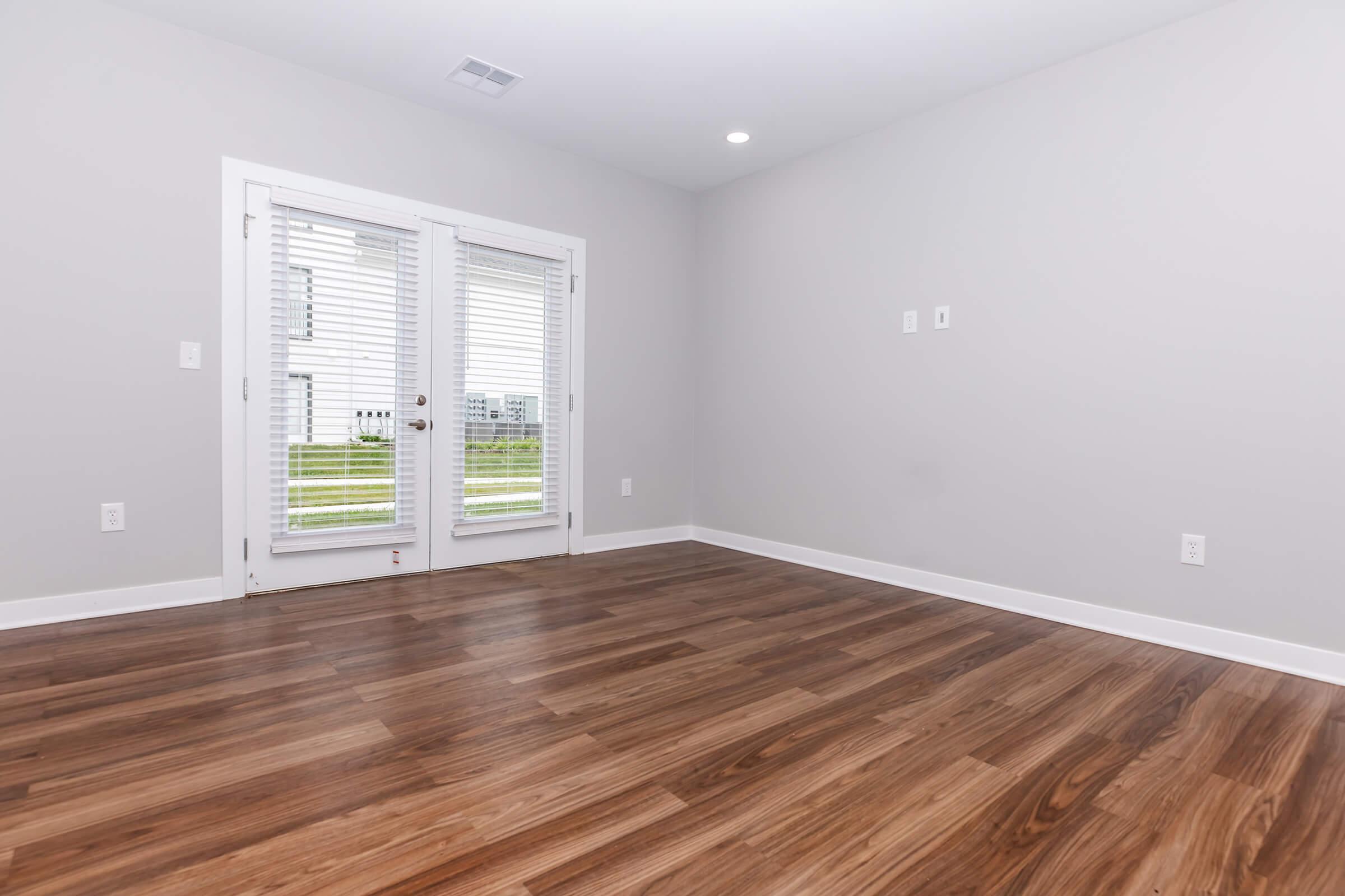 A spacious, empty room with light gray walls and hardwood flooring. It features a set of double doors with white blinds that lead to an outdoor area, providing plenty of natural light. The room has no furniture or decorations, creating an open and minimalist feel.