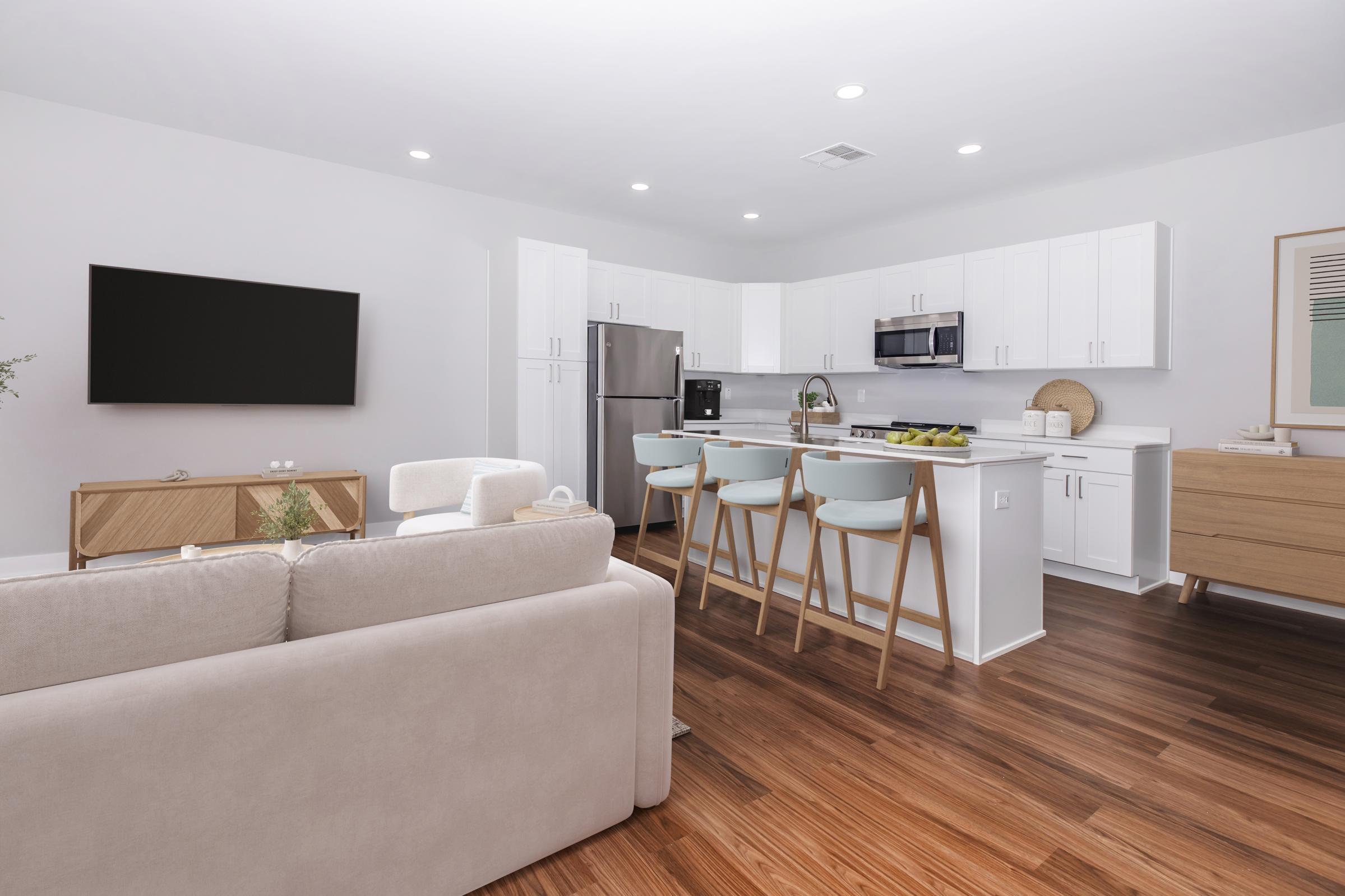 A modern kitchen and living area featuring white cabinetry, stainless steel appliances, and a large television. The kitchen island has four light blue chairs and there are wooden accents. Light wooden flooring and neutral-toned furniture create a bright, inviting atmosphere.