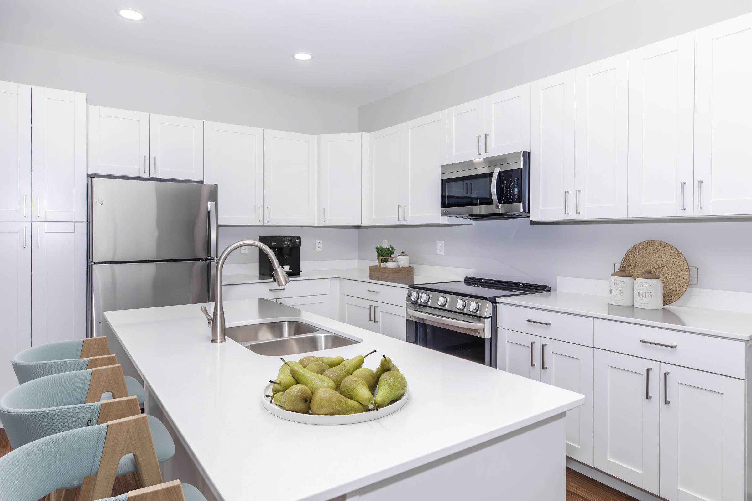 Modern kitchen featuring white cabinetry, stainless steel appliances, and a large island with a sink. A bowl of green pears is centrally placed on the island, complemented by light blue bar stools. The space is bright and inviting, showcasing a clean and contemporary design aesthetic.